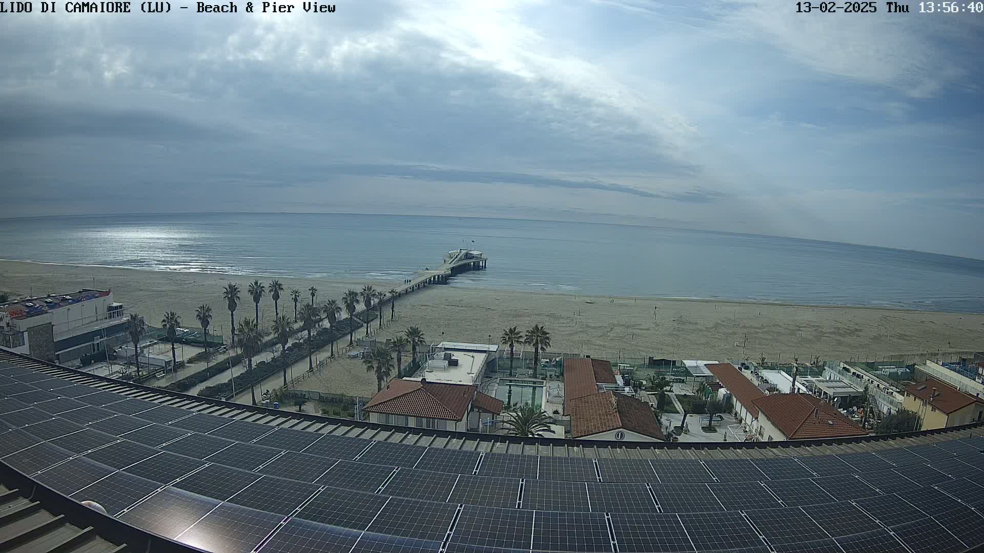 A beach with a pier, palm trees, and buildings is viewed from above a rooftop covered in solar panels under a partly cloudy sky.