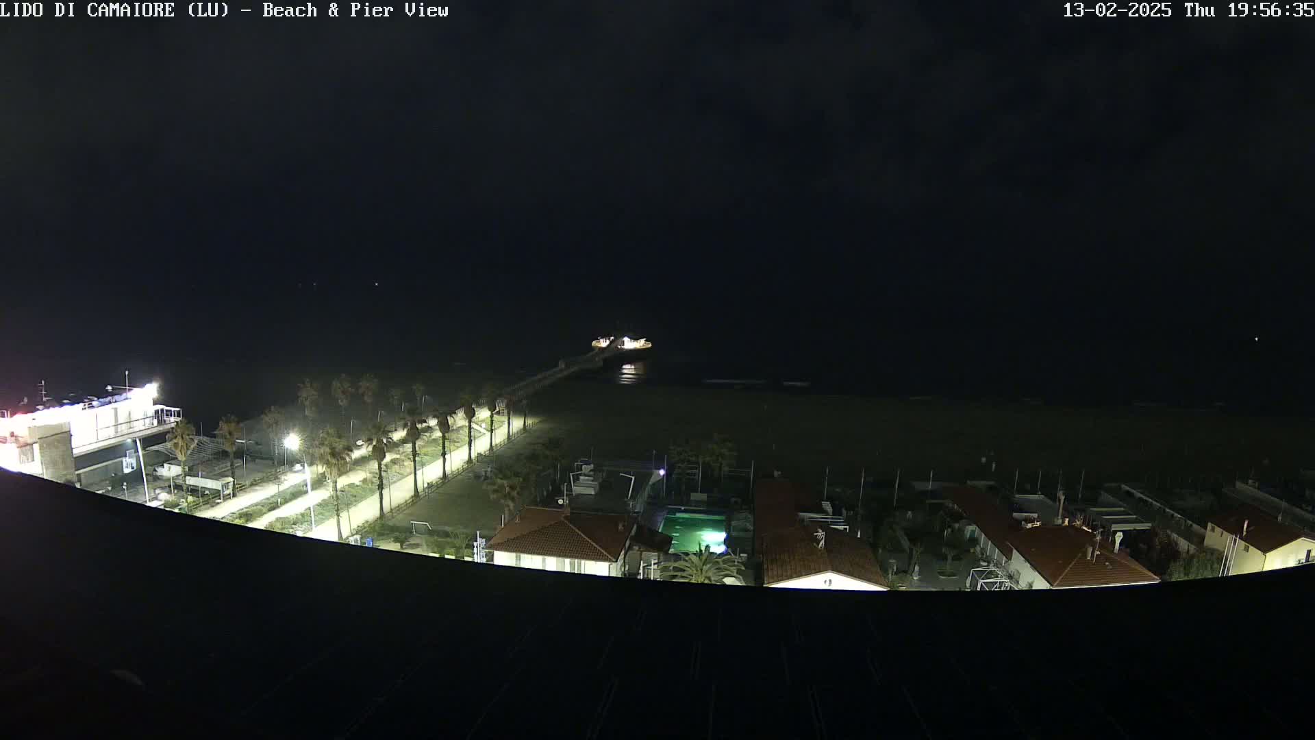 A nighttime view of a beach, pier, and buildings, under a dark, cloudy sky.