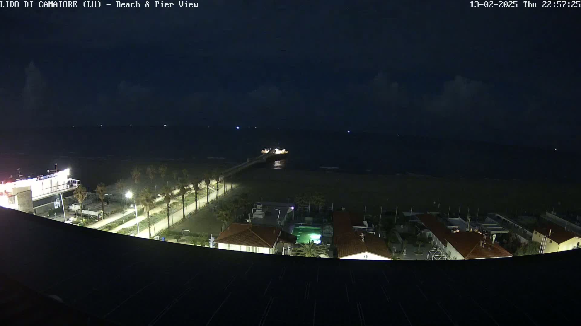 A nighttime, high-angle view shows a beach, pier, and buildings under a partly cloudy sky.