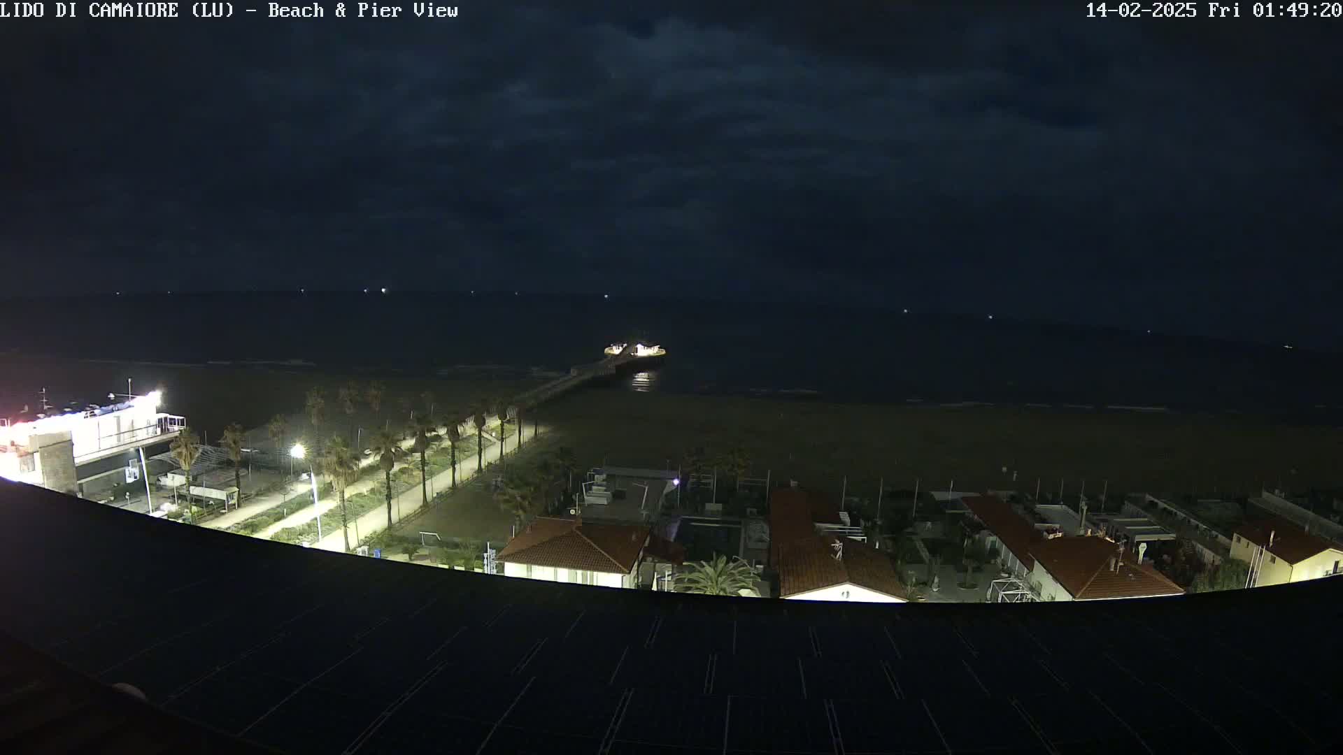 A nighttime view of a beach, pier, and coastal buildings under a cloudy sky.