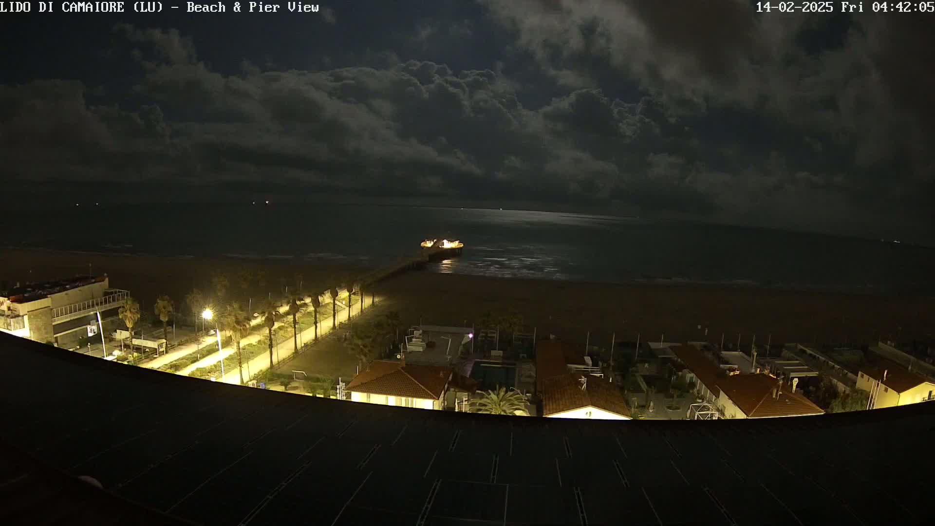 A nighttime view of a beach, pier, and coastal buildings under a partly cloudy sky.