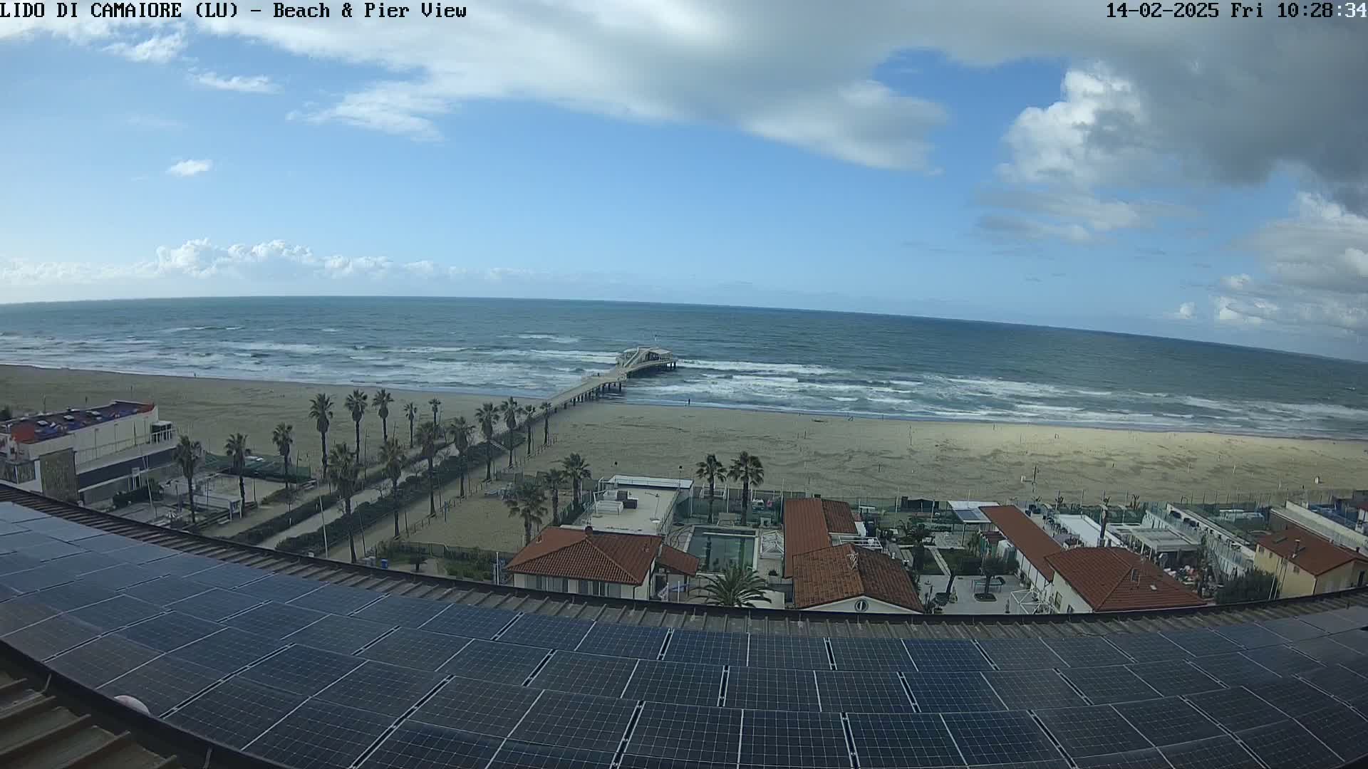 A sandy beach with a pier extends into a wavy ocean under a partly cloudy sky, viewed from above a rooftop covered in solar panels.