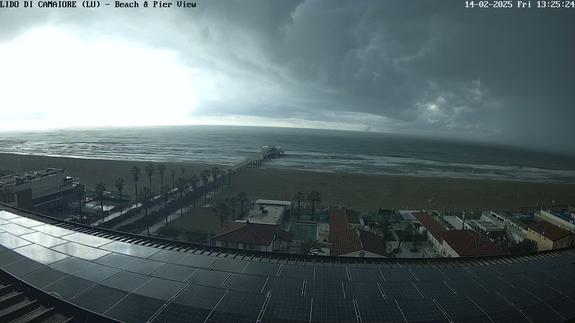 A beach with a pier is viewed from a rooftop with solar panels under a dark, cloudy sky.