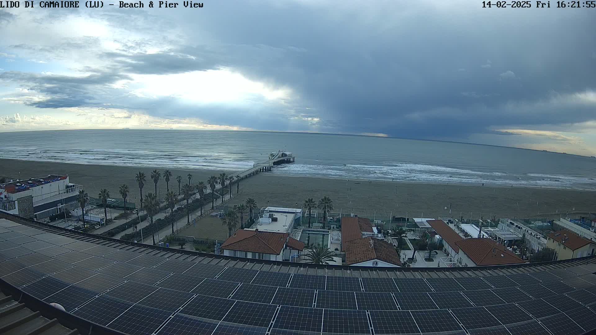 A view from a rooftop with solar panels shows a beach with a pier, palm trees, buildings, and a partly cloudy sky with dark storm clouds.