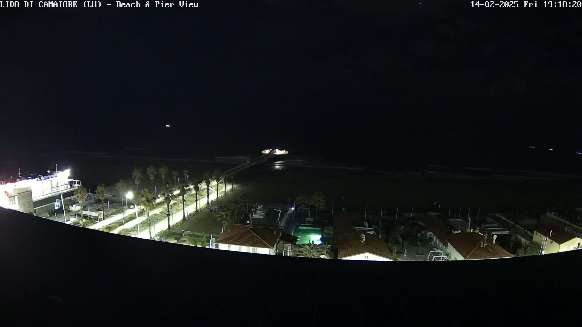 A nighttime, high-angle view of a beach, pier, and buildings illuminated by artificial light, with a dark ocean in the background.