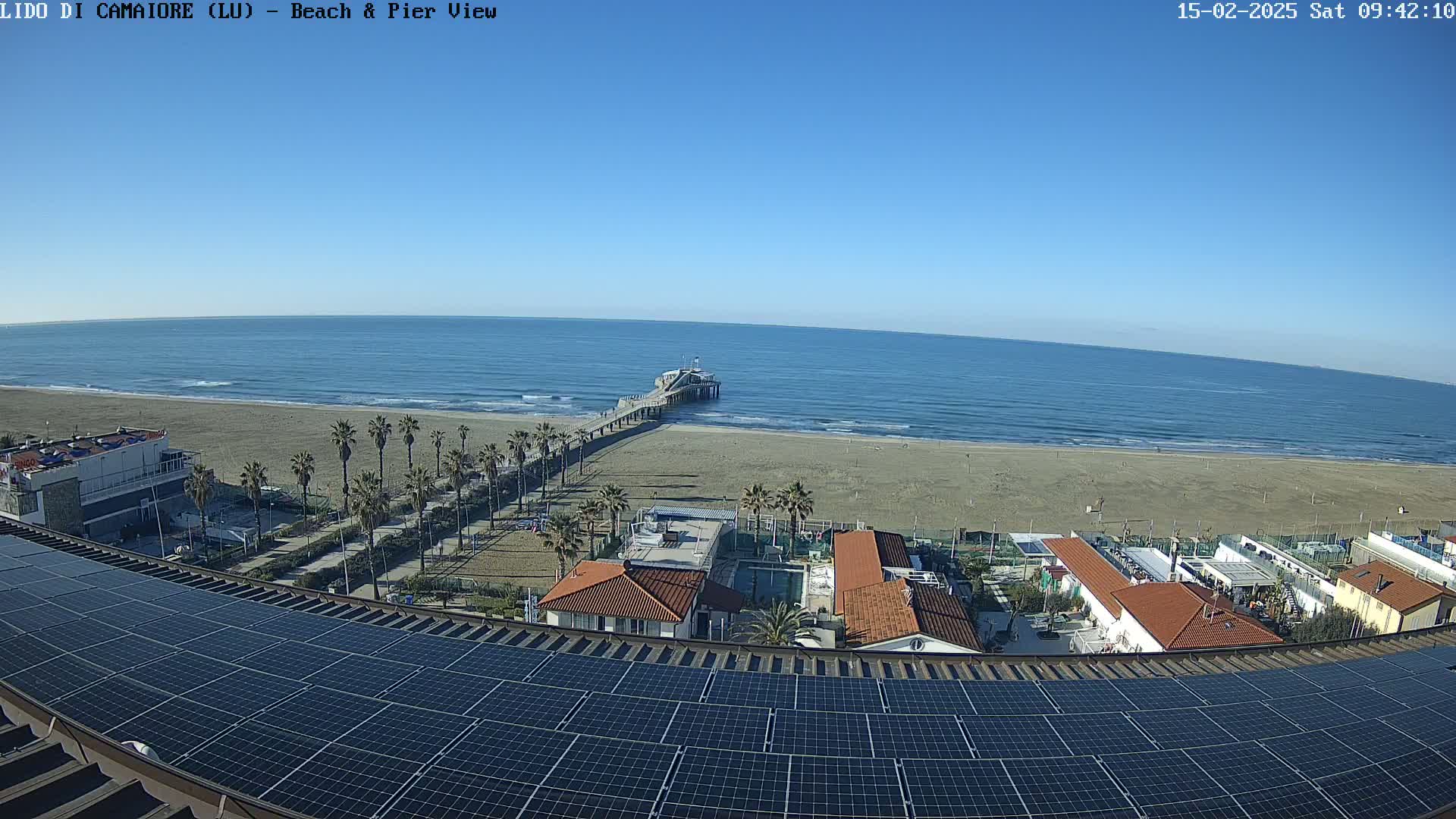 A long pier extends into a calm, blue ocean from a sandy beach, overlooked by buildings with solar panels on their roofs under a clear sky.