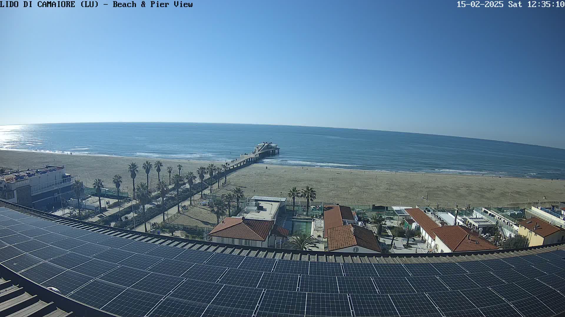 A seaside view on a sunny day shows a pier extending from a sandy beach into a calm ocean, with palm trees, buildings, and solar panels visible in the foreground.
