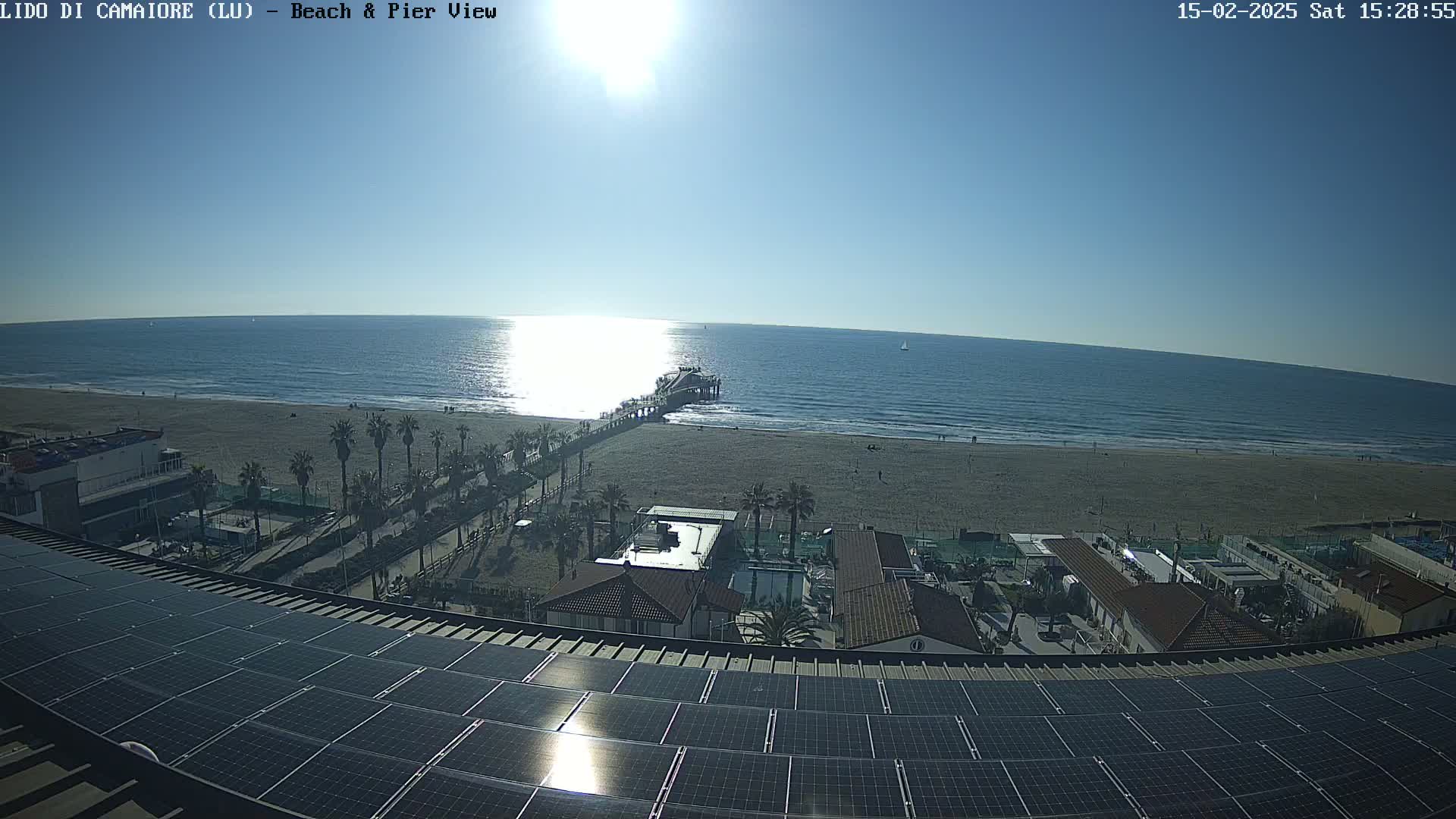 A sunny day reveals a beach scene with a pier extending into a calm ocean, palm trees lining the shore, and a rooftop covered in solar panels overlooking the scene.