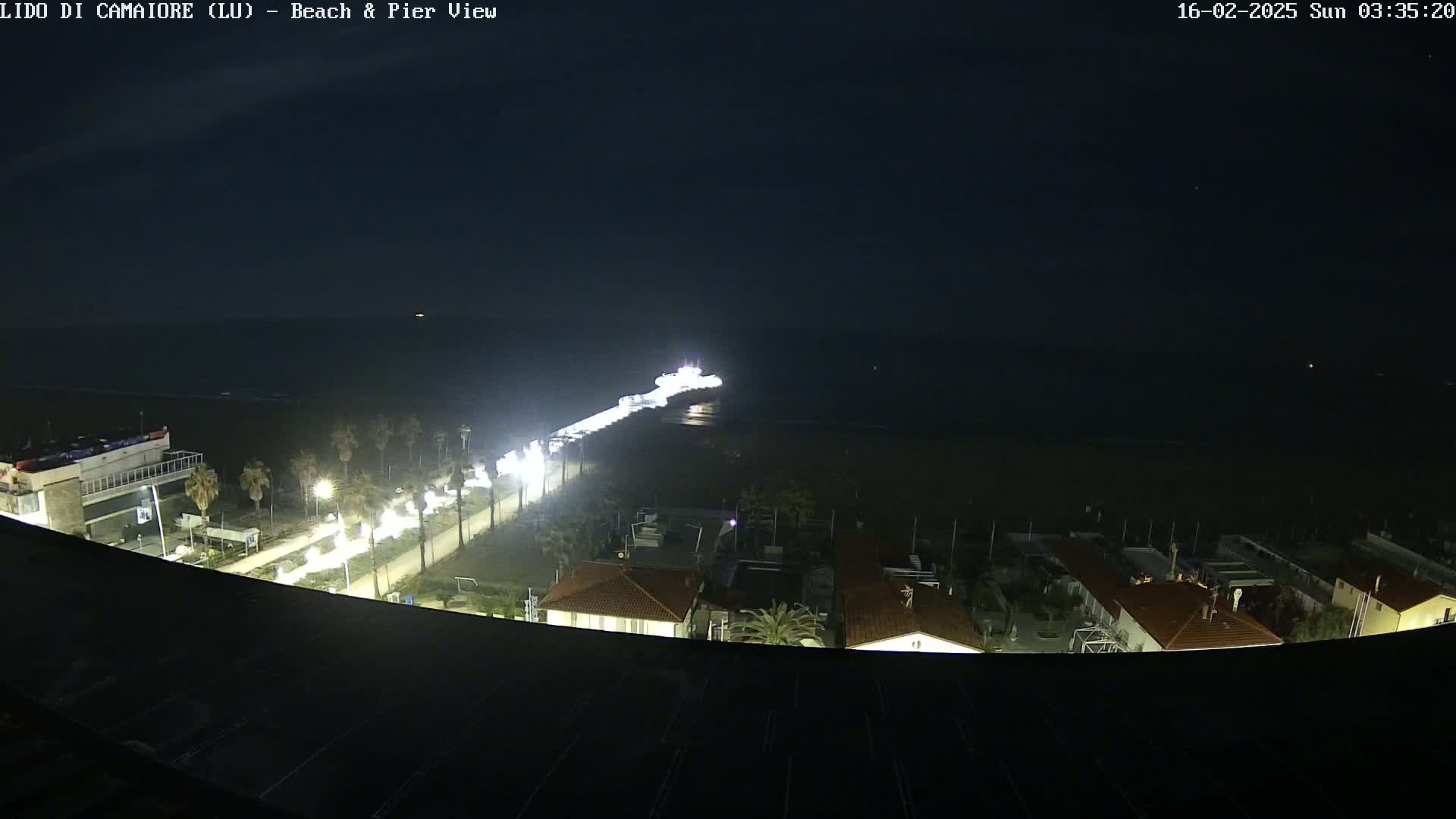 A nighttime, aerial view shows a pier extending into a dark ocean, with a brightly lit beachfront road and buildings visible below.