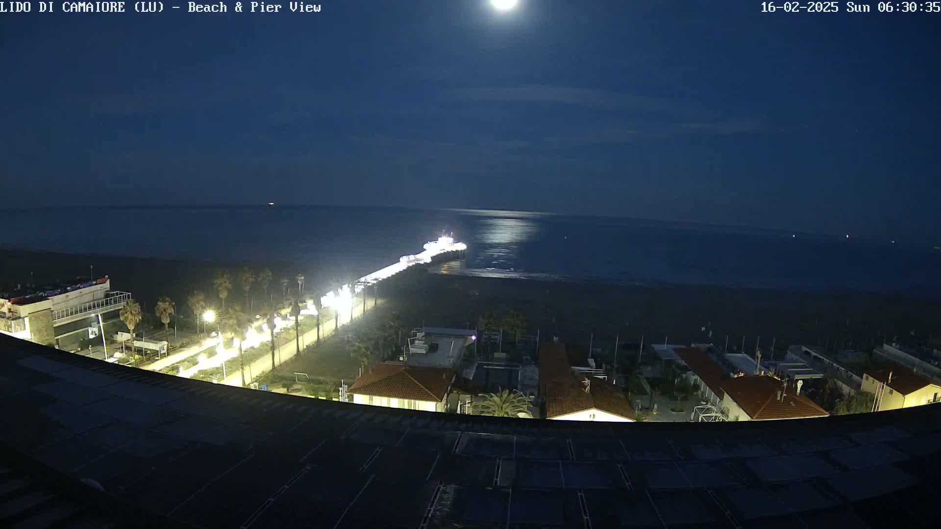 A brightly lit pier extends from a moonlit beach at night, with buildings and streetlights visible along the shoreline.