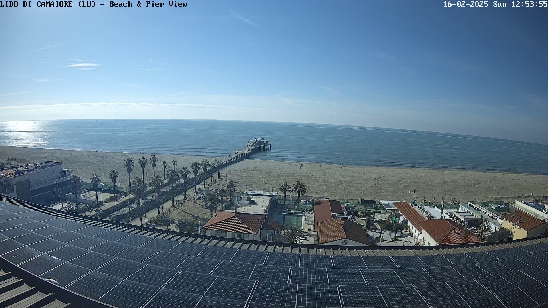 A beach with a pier, buildings, palm trees, and a solar panel array on a rooftop are visible under a clear, sunny sky.