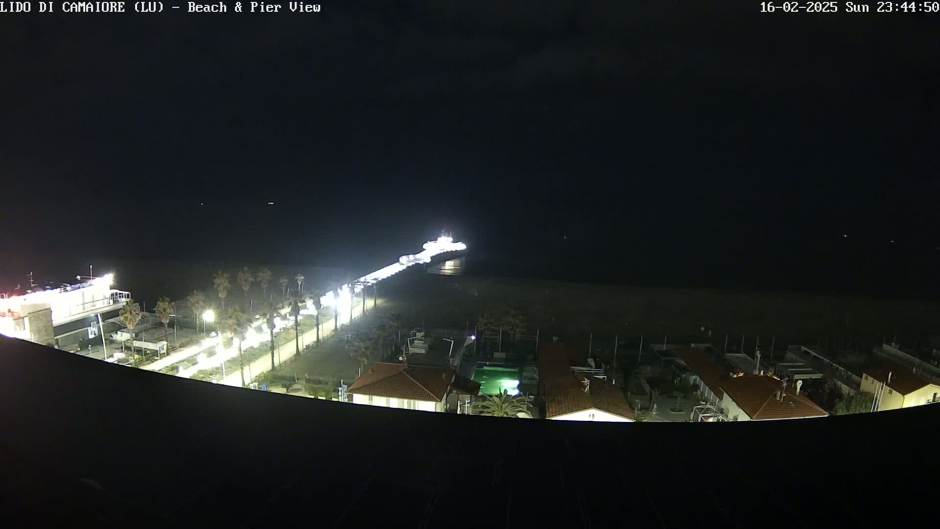 A nighttime view of a beach and pier with a boat at the end of the pier,  illuminated by lights along a walkway and buildings nearby.
