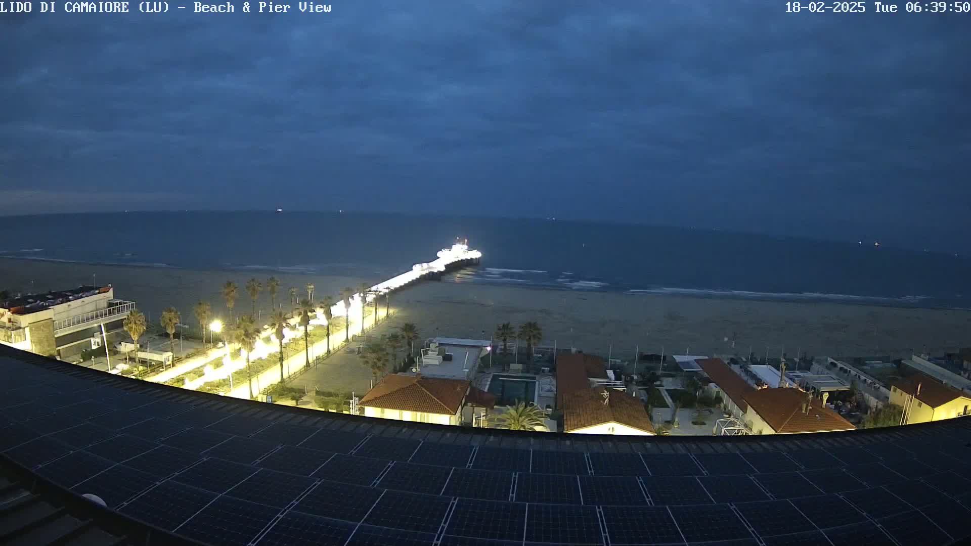 A nighttime beach scene shows a pier extending into a calm sea under a dark, cloudy sky, with illuminated buildings and palm trees along the beachfront, all viewed from a rooftop with solar panels.