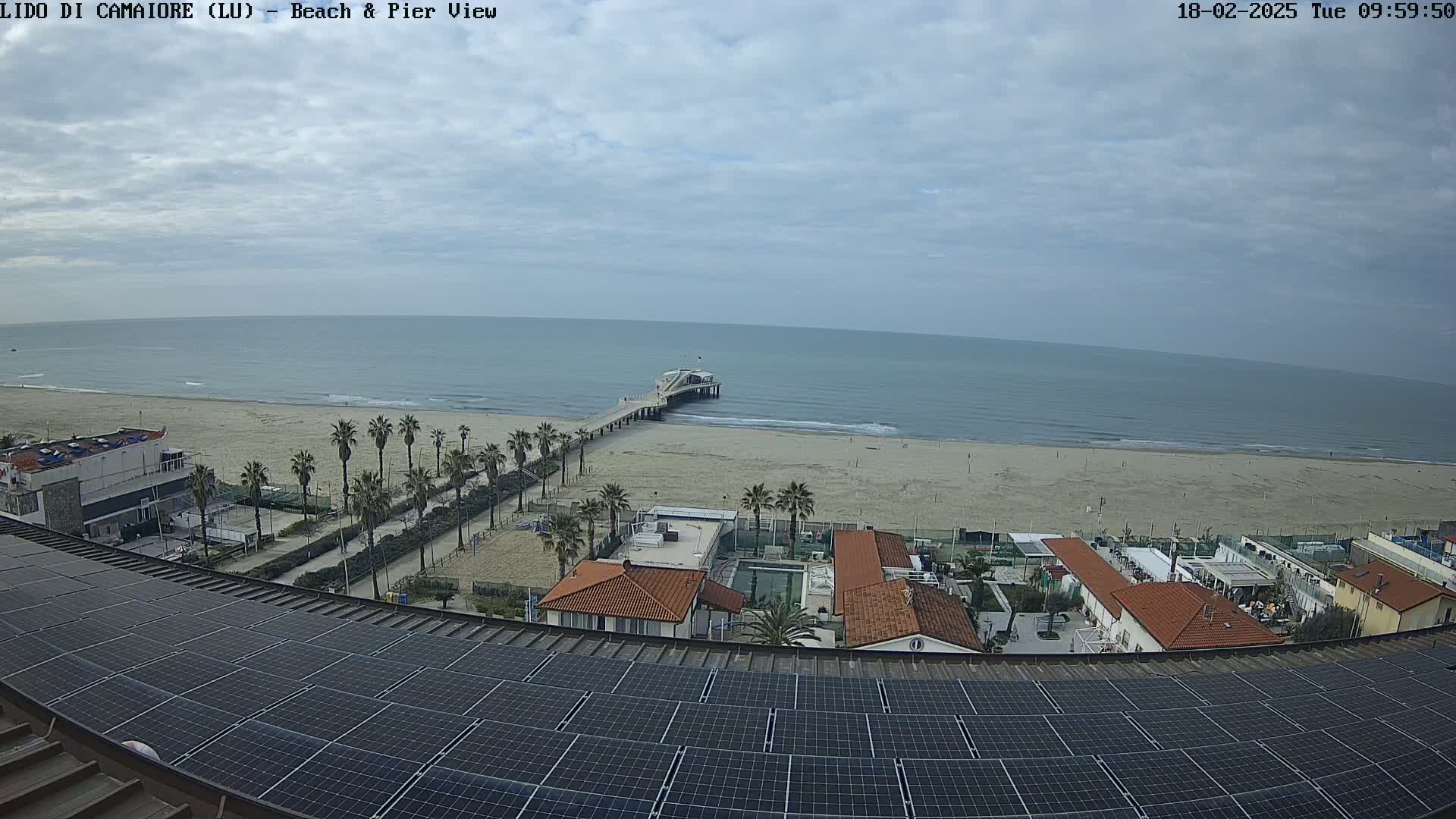 A beach with a pier extends into a calm ocean under a mostly cloudy sky, viewed from above a rooftop covered in solar panels.