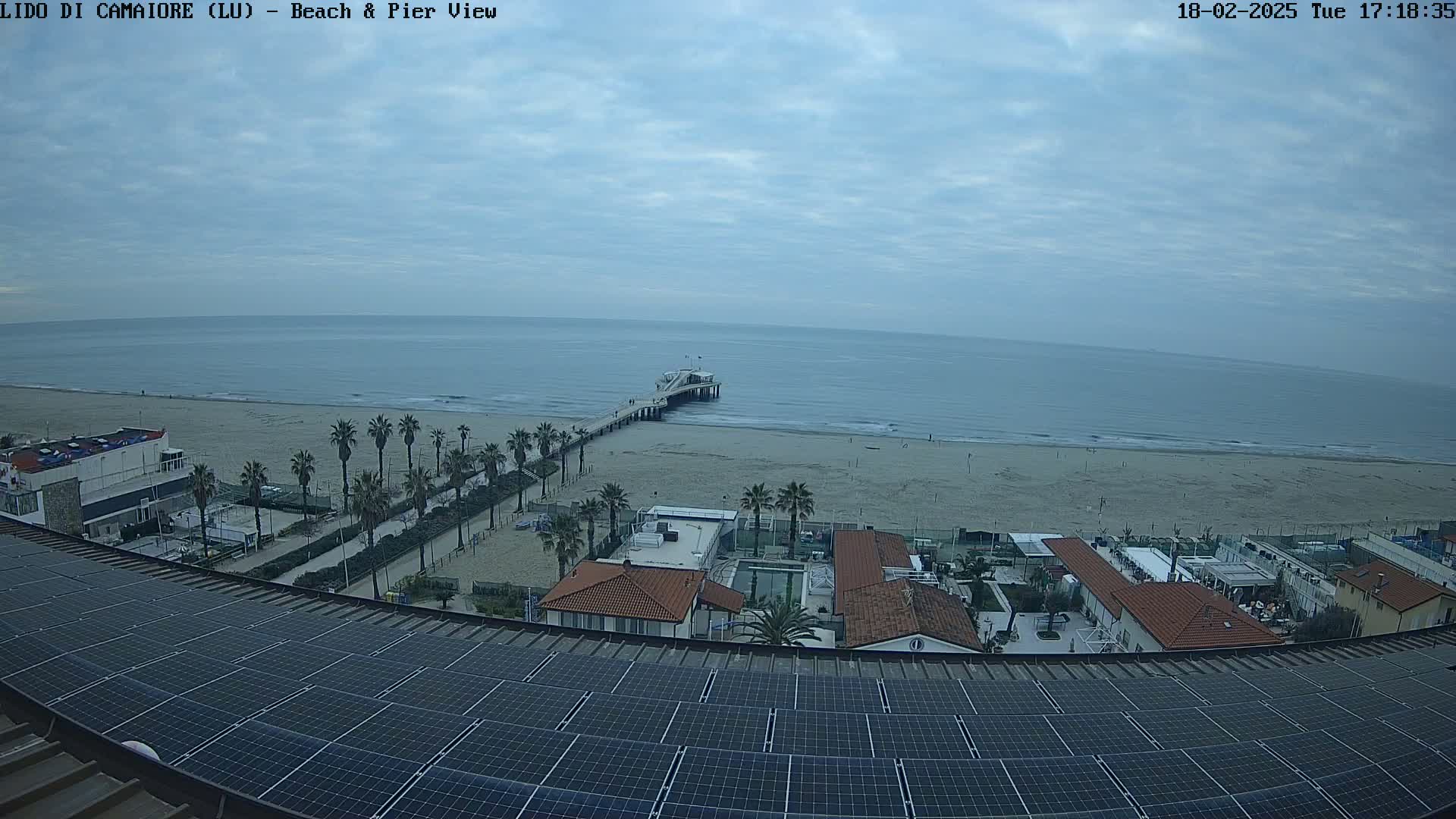 An overcast sky overlooks a beach with a pier, palm trees, and buildings, all viewed from above a rooftop covered in solar panels.