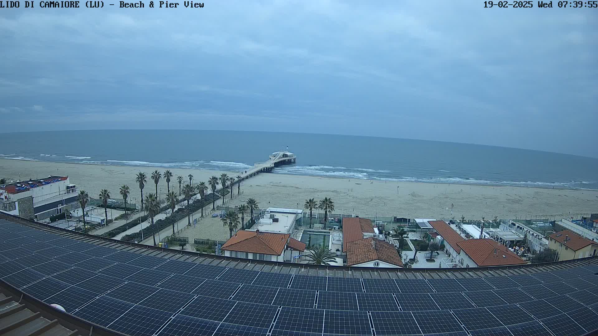 A beach with a pier, palm trees, buildings, and a solar panel array on a rooftop are visible under a cloudy sky.