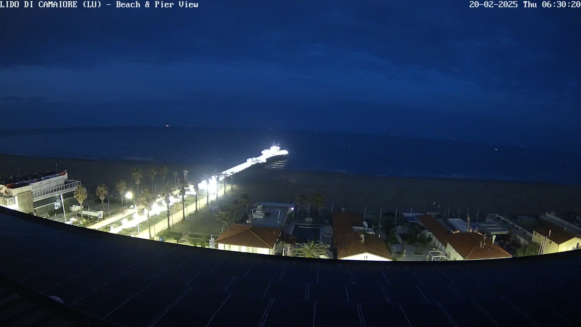 A nighttime view of a beach with a pier extending into a dark blue sea, under a mostly clear sky, showing illuminated buildings and palm trees along the shore.