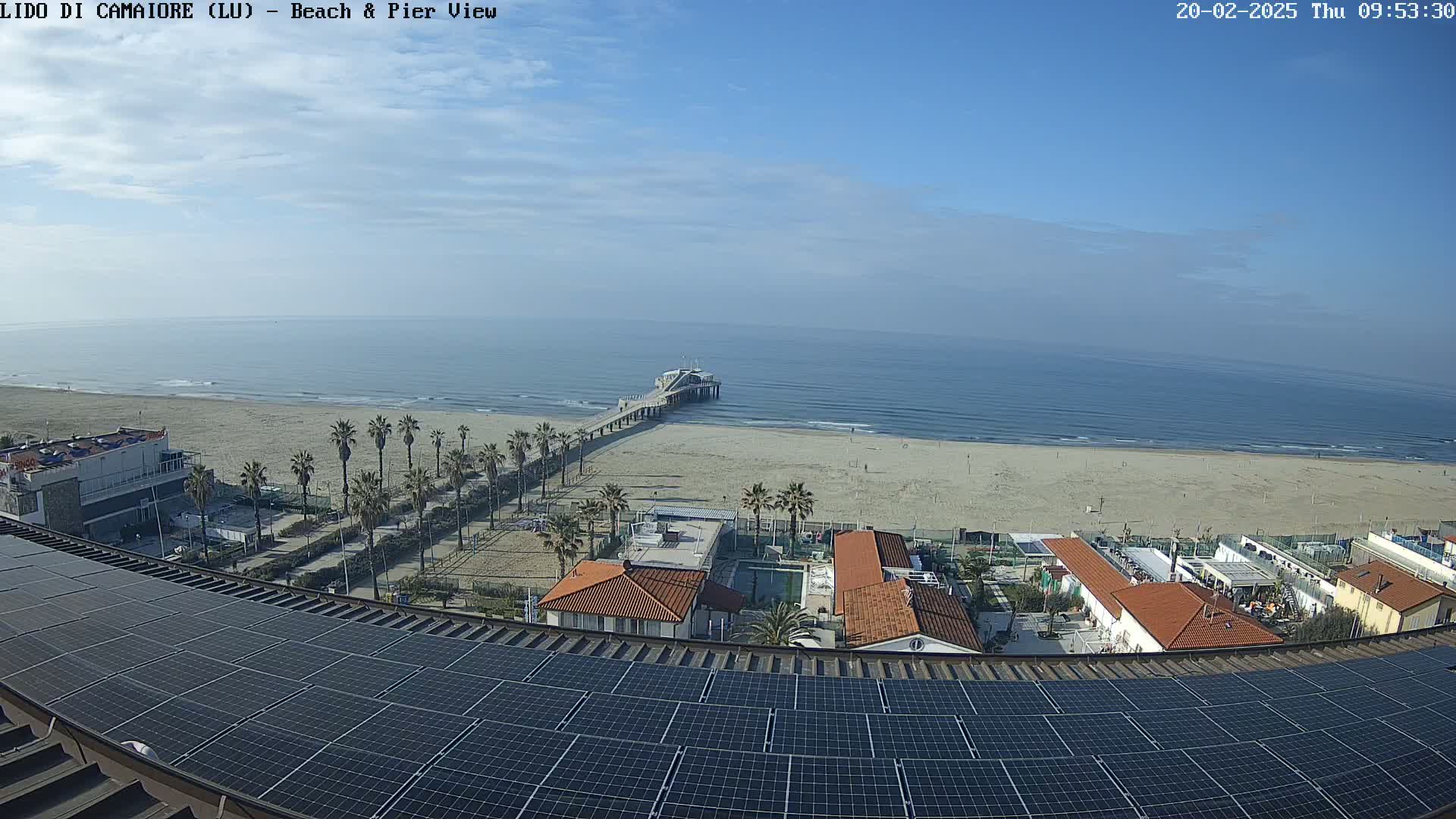 A clear day reveals a sandy beach, a long pier extending into a calm sea, beachfront buildings, and palm trees, all viewed from a rooftop covered in solar panels.