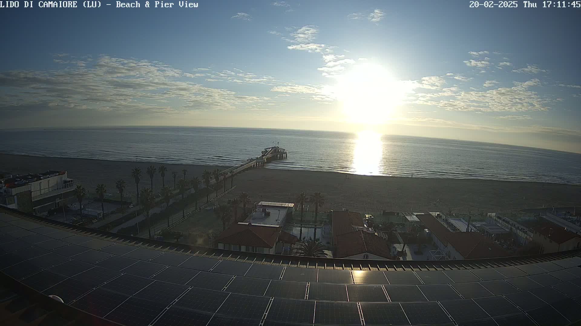 A beach with a pier is visible from an elevated vantage point showing a mostly sunny sky with some clouds, palm trees lining a sandy shore, and buildings with solar panels on a roof.