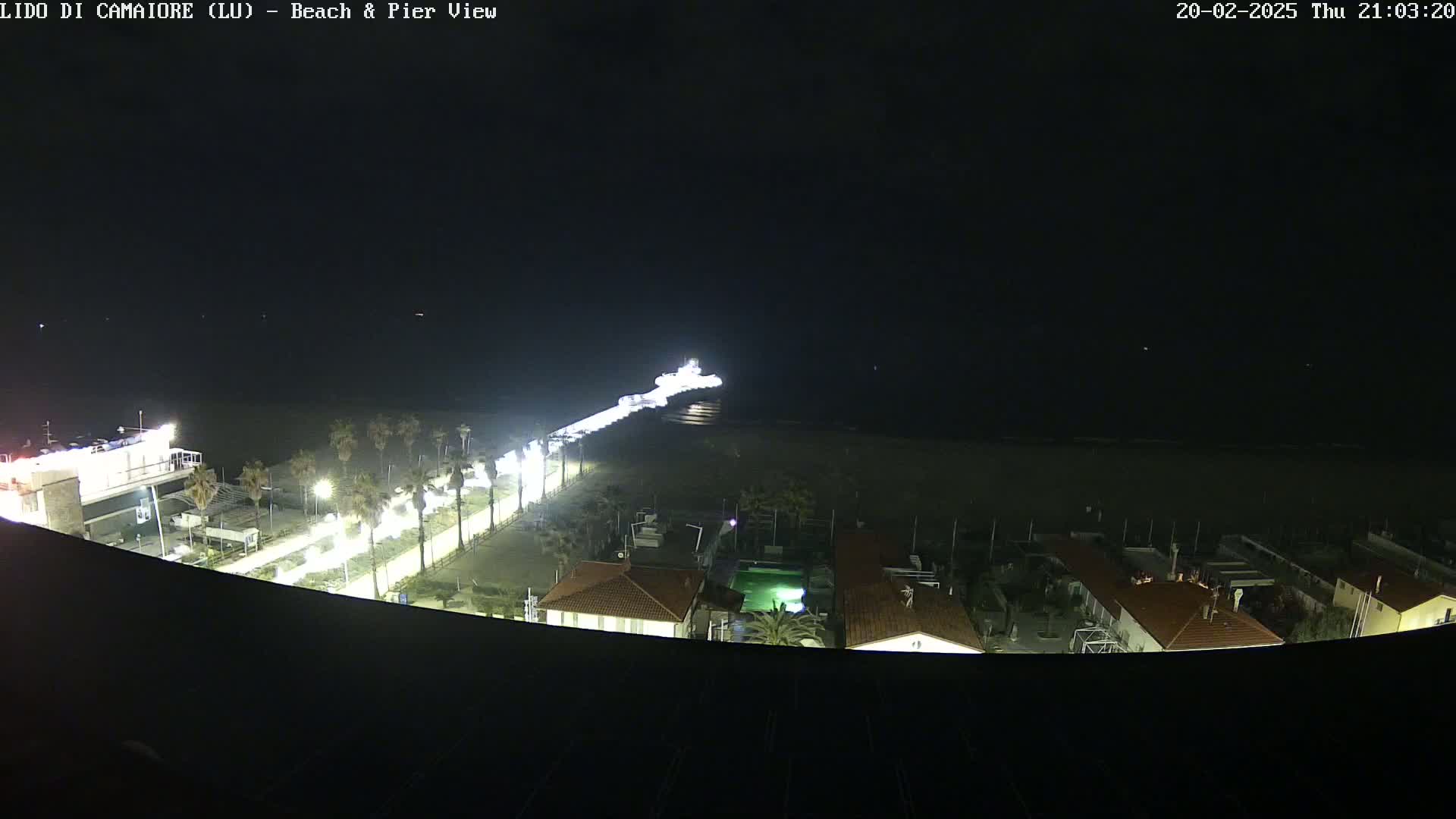 A nighttime, high-angle view shows a pier extending into a dark ocean, with a brightly lit beach and beachfront buildings below.
