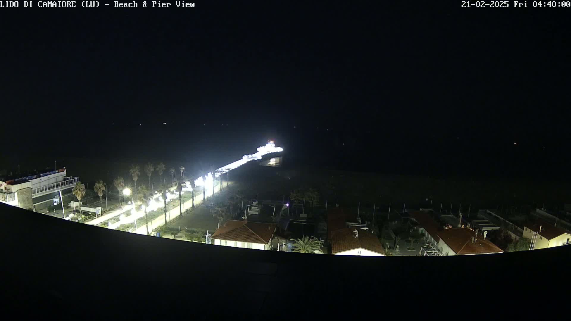 A nighttime, aerial view of a pier extending from a beach with buildings and palm trees illuminated along the shore.