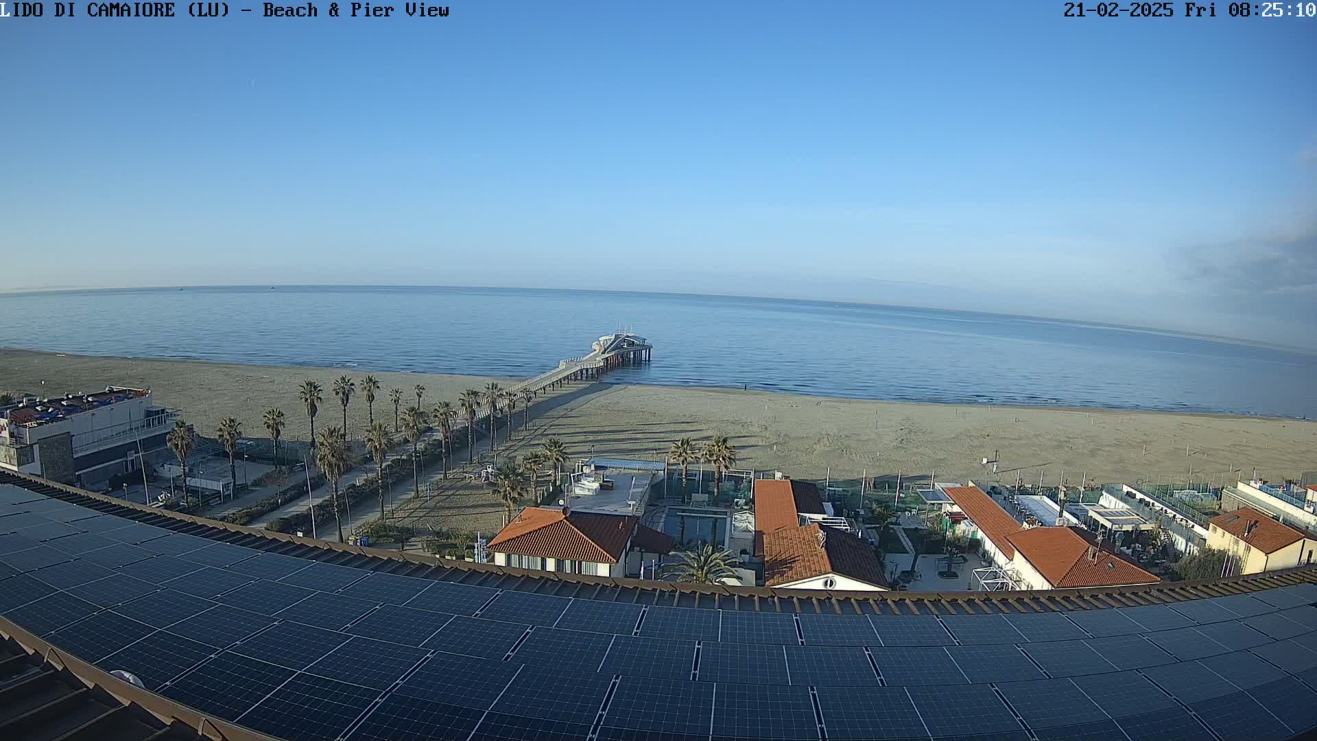 A long pier extends into a calm, blue ocean from a sandy beach lined with palm trees, all viewed from above a building with solar panels on its roof.