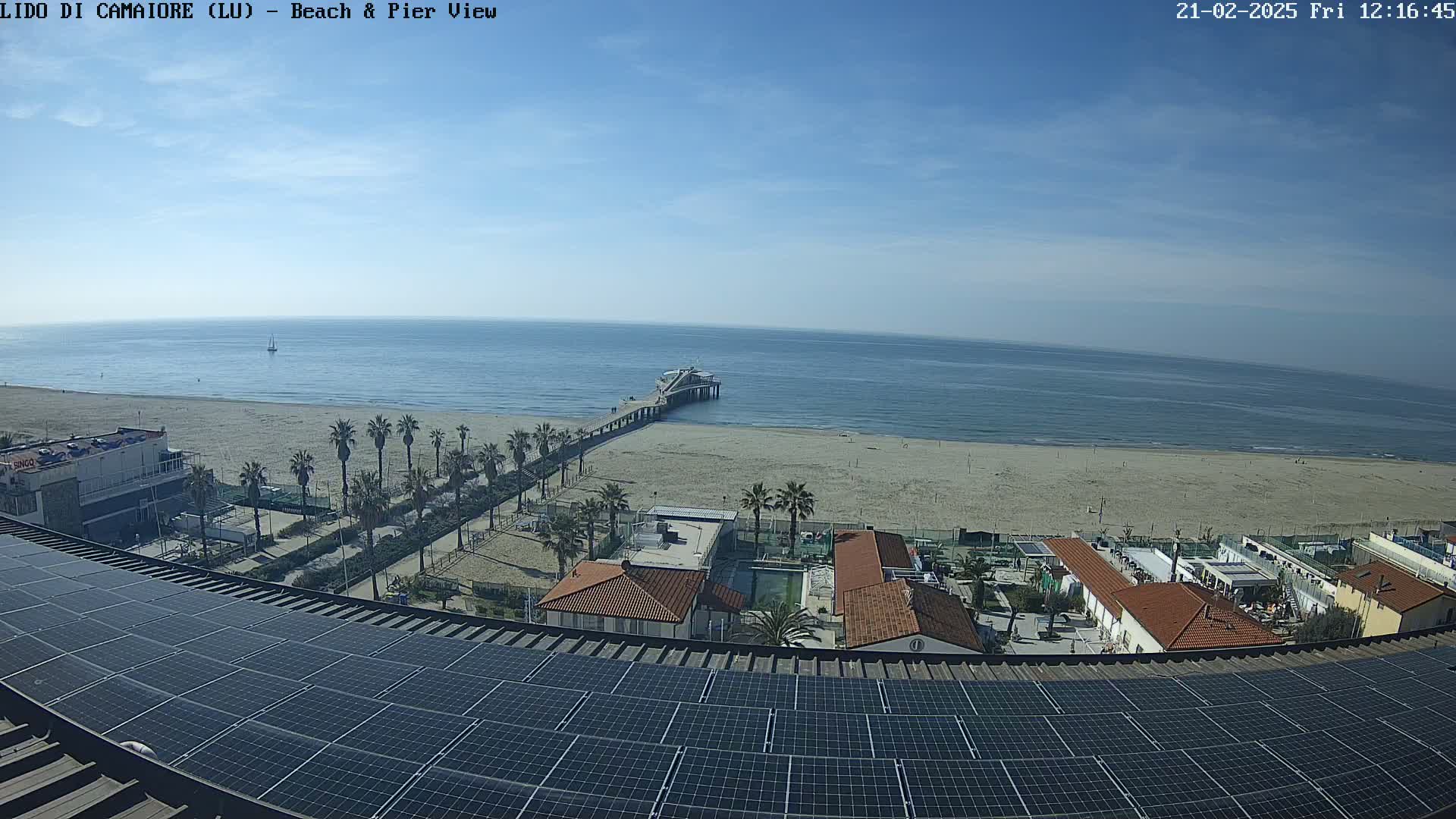 A beach with a pier extends into a calm ocean under a clear blue sky, viewed from a rooftop covered in solar panels.