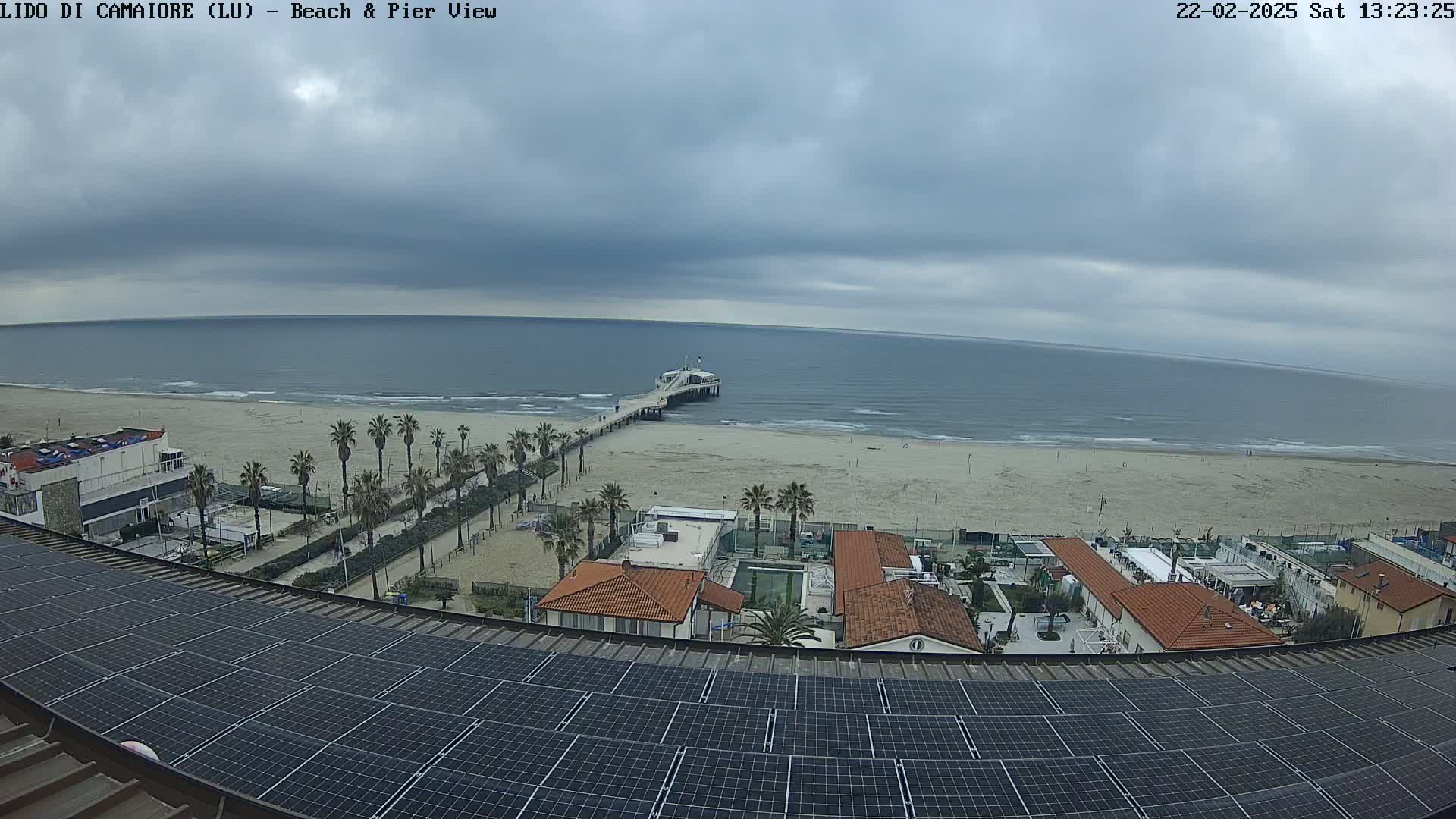 An overcast day reveals a beach scene with a pier extending into a calm sea, viewed from a rooftop with solar panels.