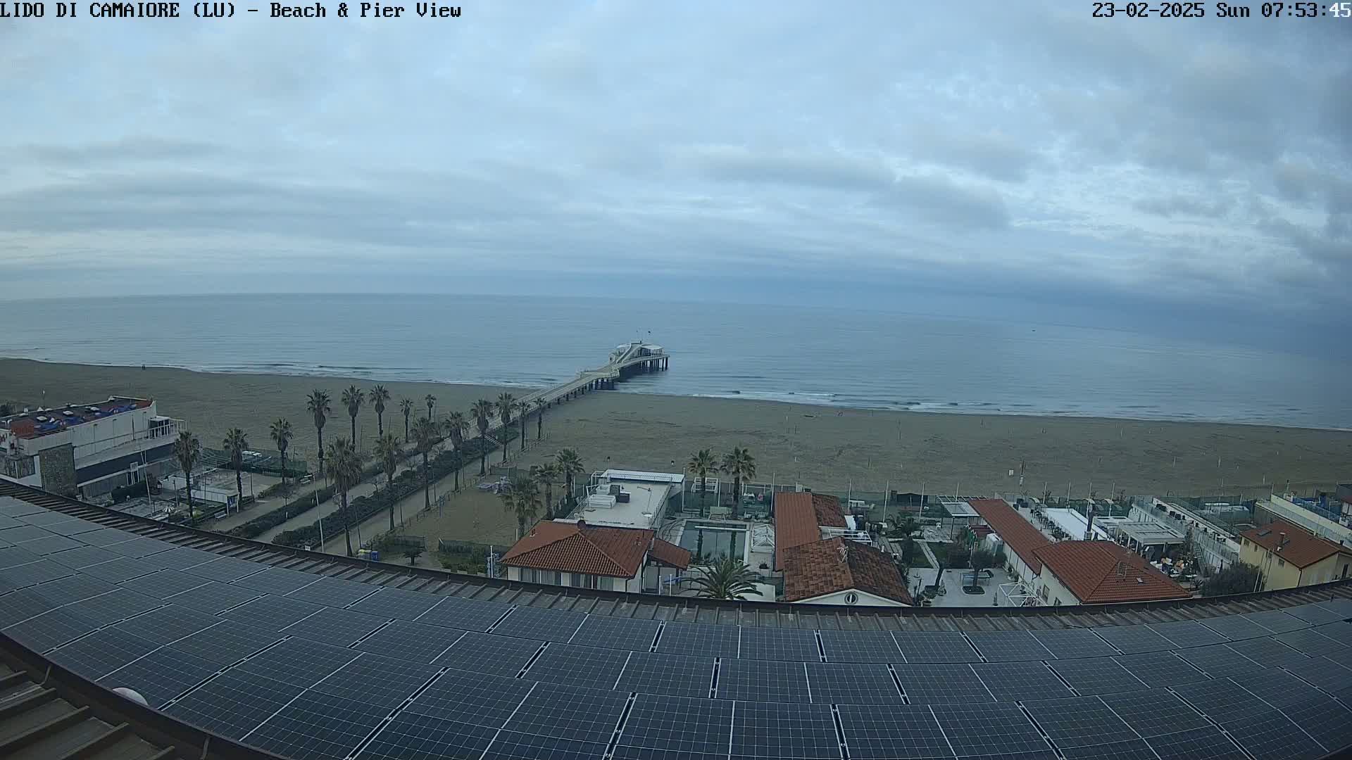 A long pier extends from a sandy beach into a calm, overcast ocean, viewed from a rooftop with solar panels.