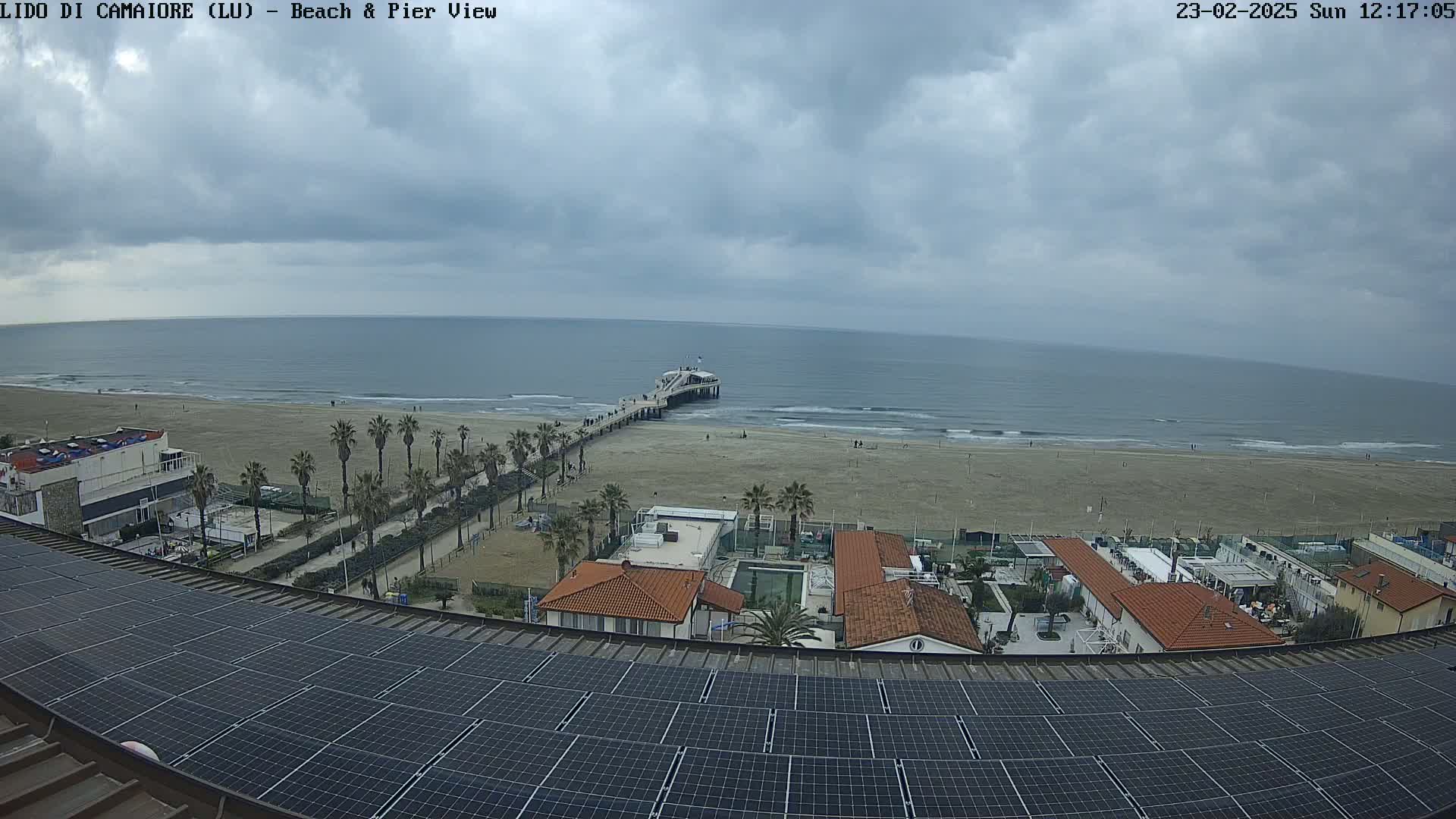 A beach with a pier, palm trees, and buildings is seen from above on a cloudy day, with solar panels in the foreground.