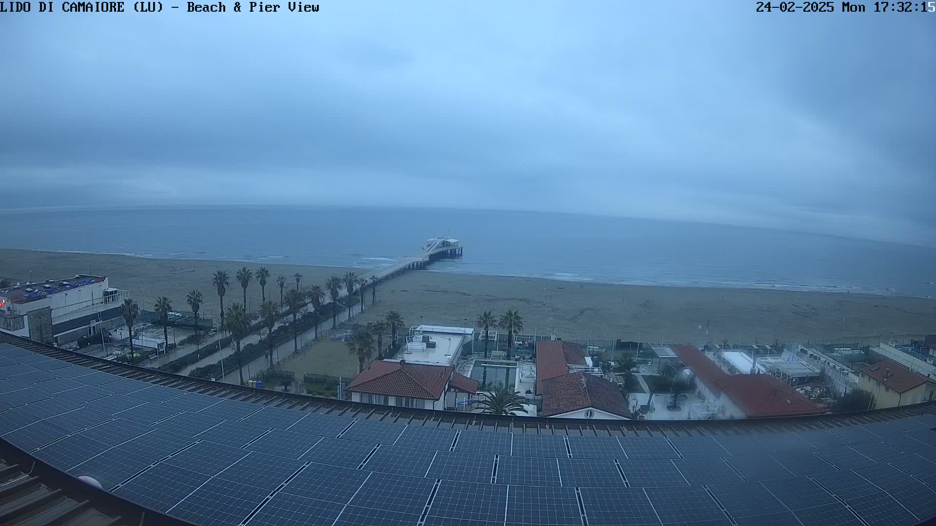 A high-angle, long shot shows a beach with a pier under an overcast sky, viewed from a rooftop with solar panels.