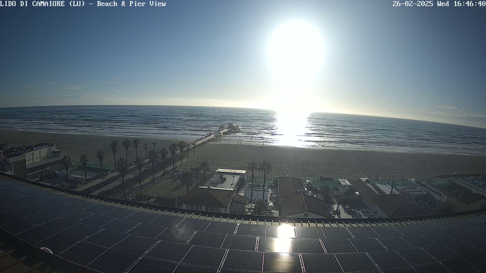 A sunny day at a beach shows a pier extending into the ocean, with palm trees and buildings visible from an elevated viewpoint, overlooking a large solar panel array.