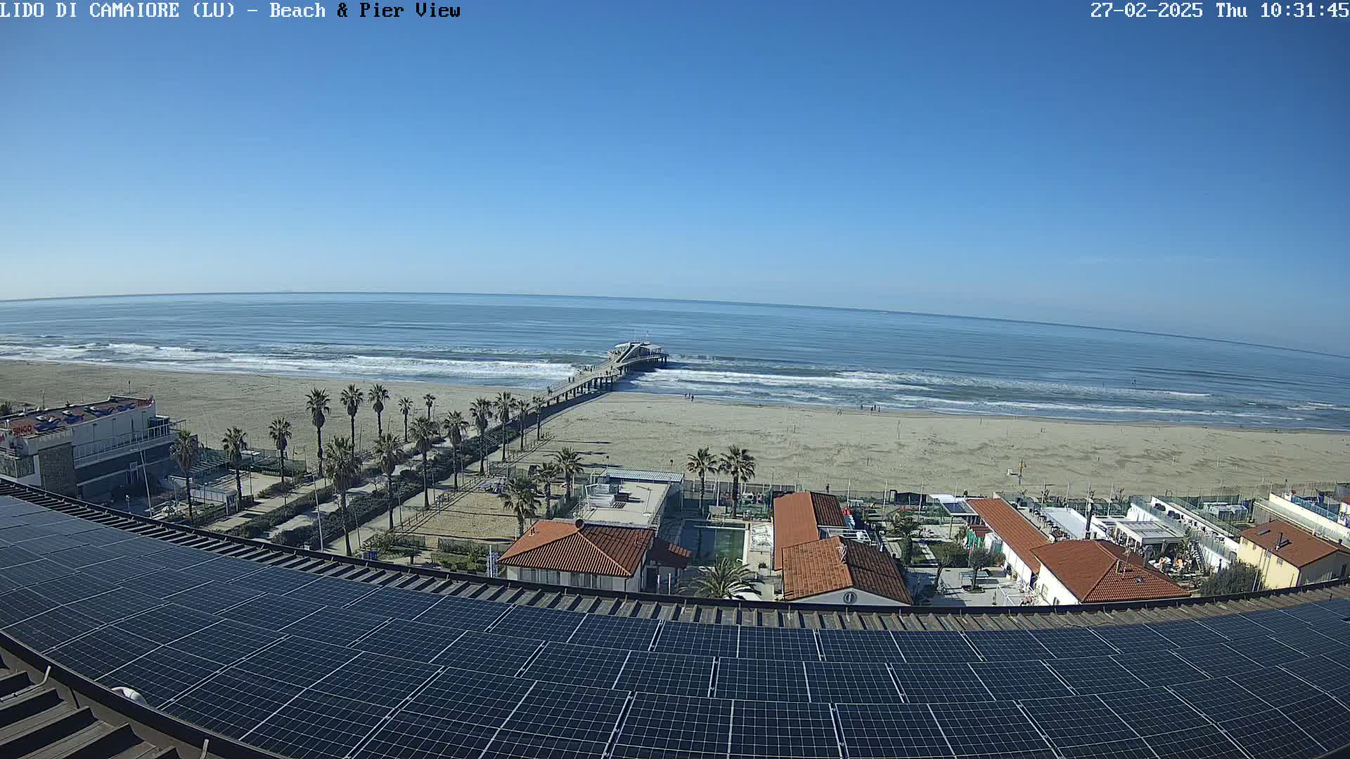 A sandy beach with a pier extends into a calm ocean under a clear blue sky, viewed from above a rooftop covered in solar panels.