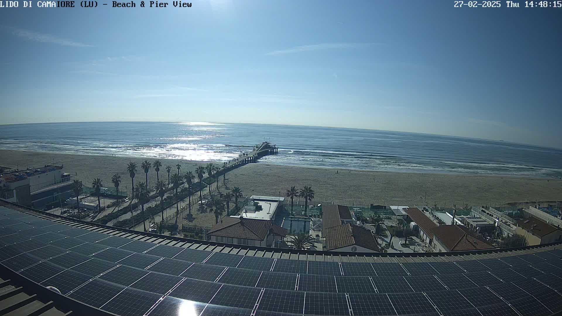 A beach with a pier extends into a calm ocean under a clear, sunny sky, viewed from a rooftop covered in solar panels.
