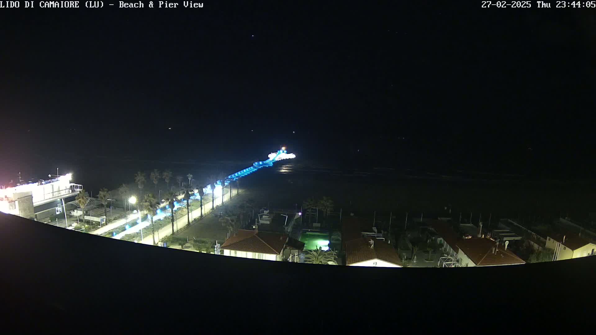 A nighttime view of a pier extending from a beach into a dark ocean, with lights illuminating the pier and the beachfront promenade.