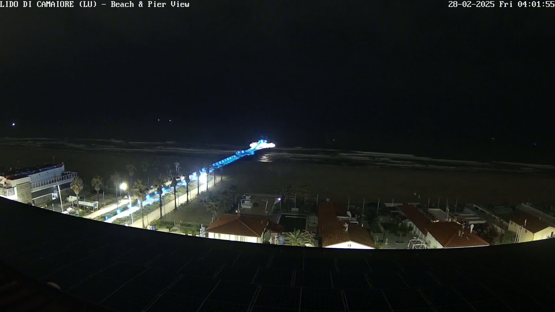 A nighttime, overhead view of a beach at night shows a pier lit with blue lights extending from the shore into the ocean, with buildings and streetlights visible along the coast.
