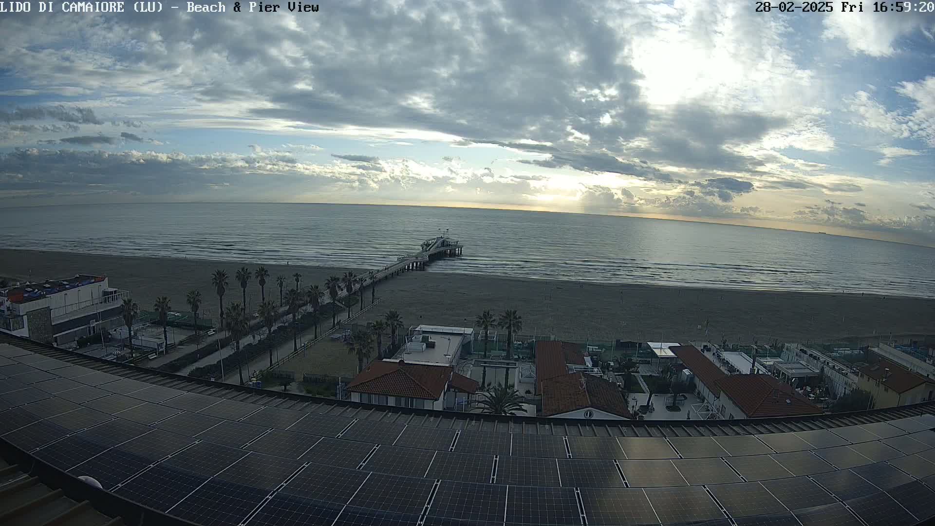 A mostly cloudy sky overlooks a beach with a pier, palm trees, buildings, and a large solar panel array on a rooftop.