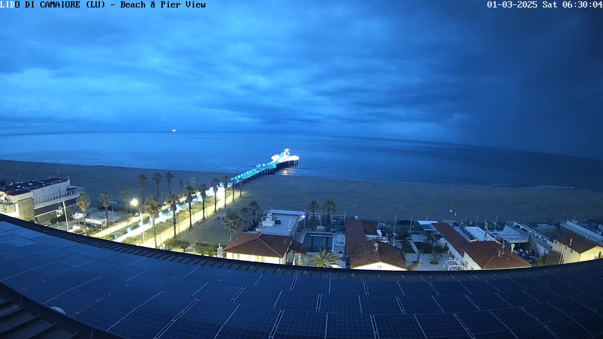 A nighttime coastal scene shows a pier extending into a calm, dark-blue sea under a cloudy sky, with a beach, palm trees, buildings, and solar panels visible in the foreground.