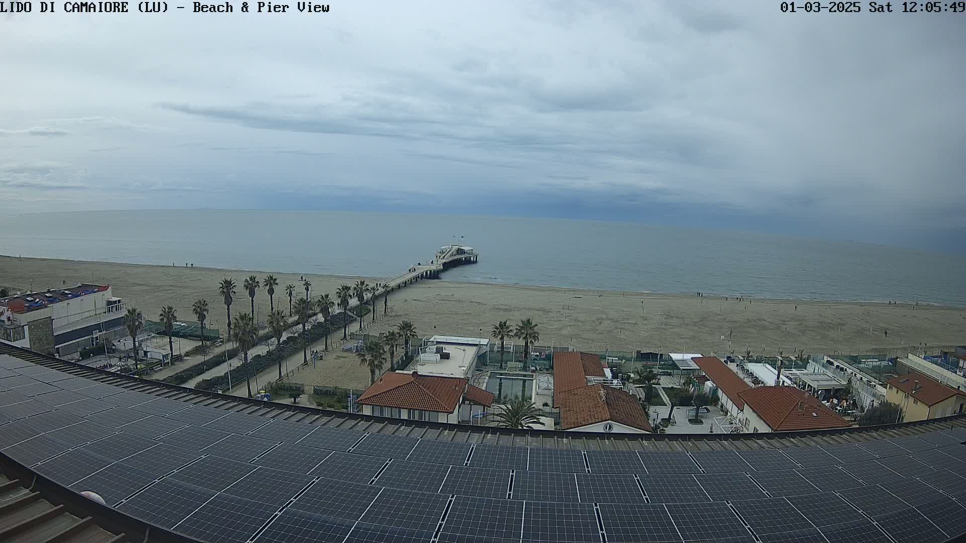 A beach with a pier extends into a calm, overcast sea, viewed from above a building equipped with solar panels.