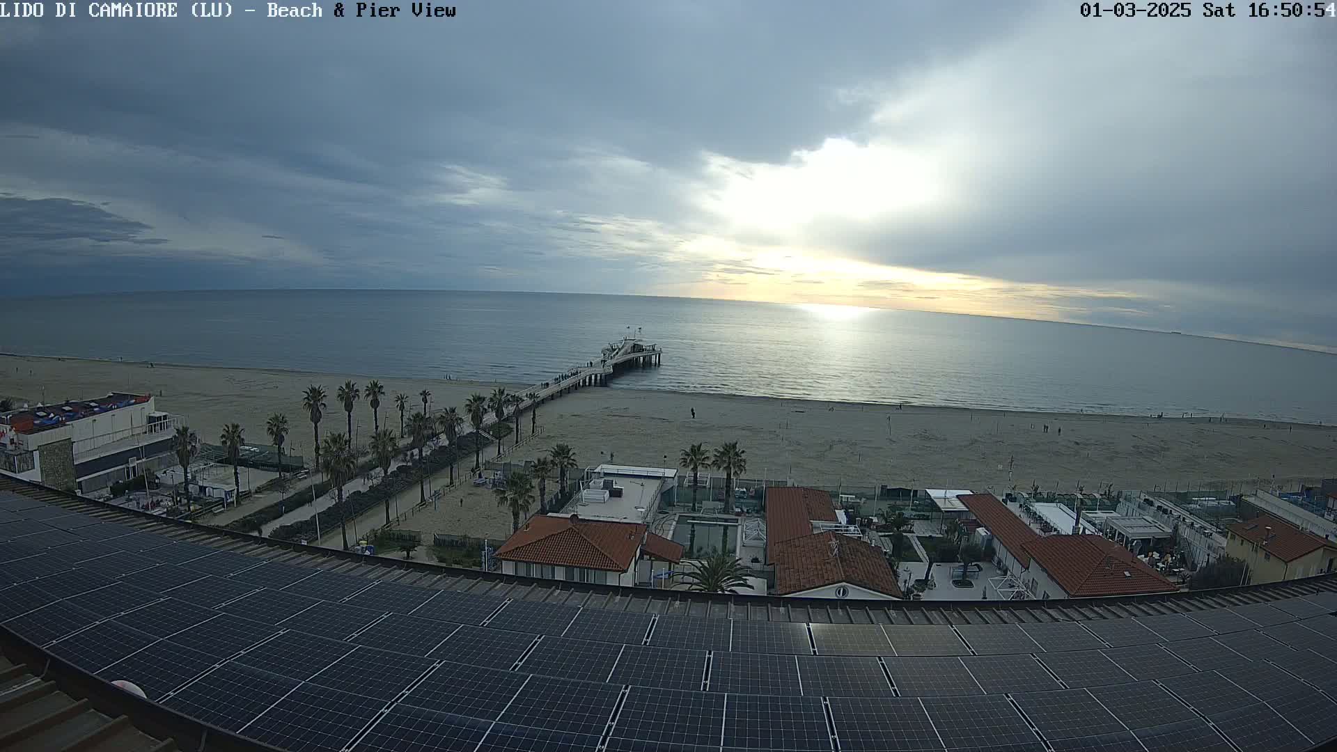 A beach with a pier and buildings is seen from above under a partly cloudy sky, with a large solar panel array in the foreground.
