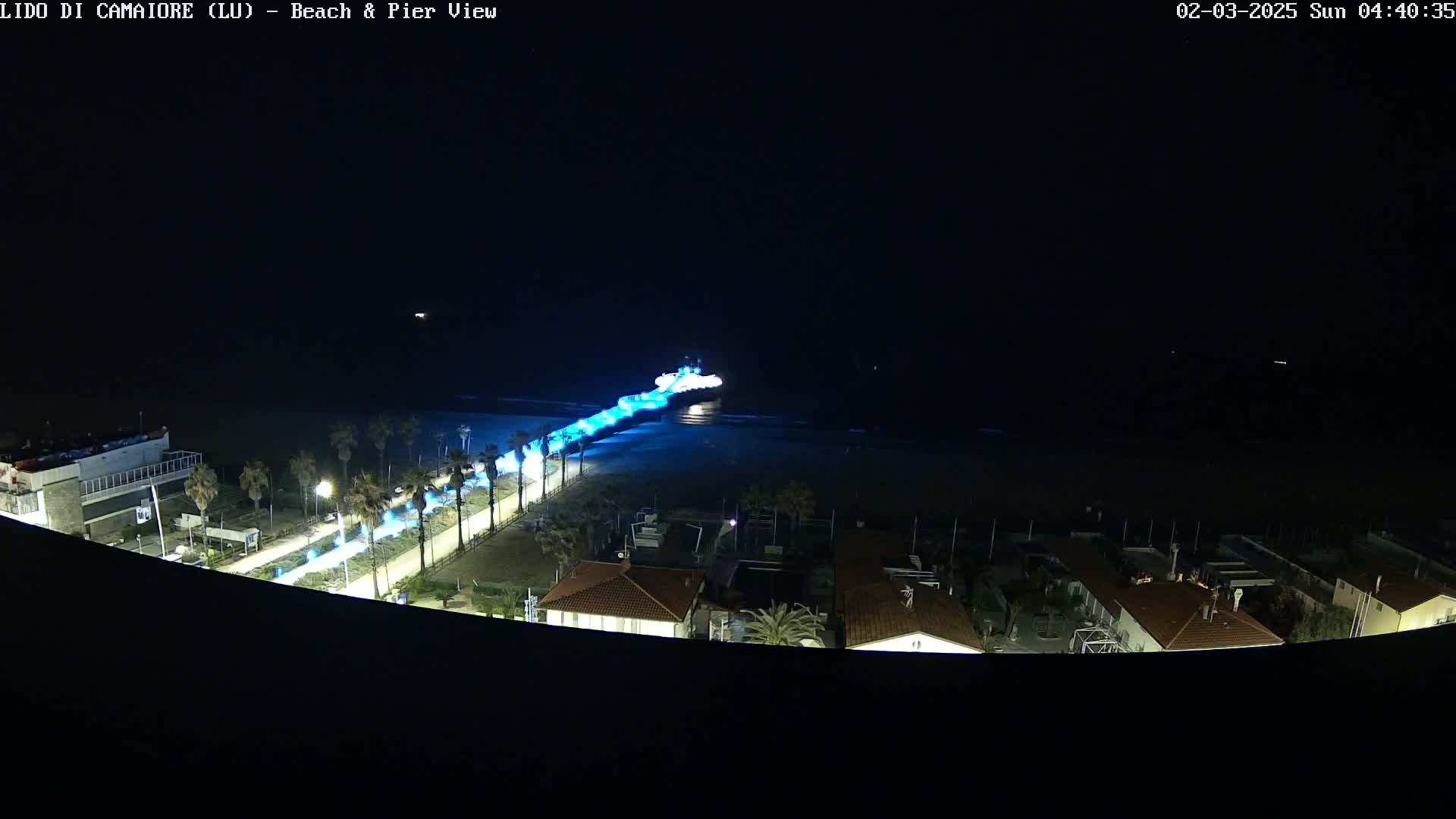 A nighttime, high-angle view shows a pier extending from a beach into dark water, illuminated by blue lights, with buildings and palm trees visible on the shore.