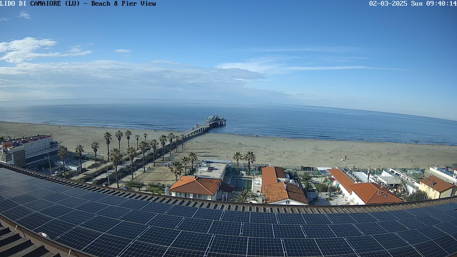 A mostly sunny day reveals a beach scene with a pier extending into a calm ocean, viewed from a rooftop covered in solar panels overlooking buildings and palm trees.