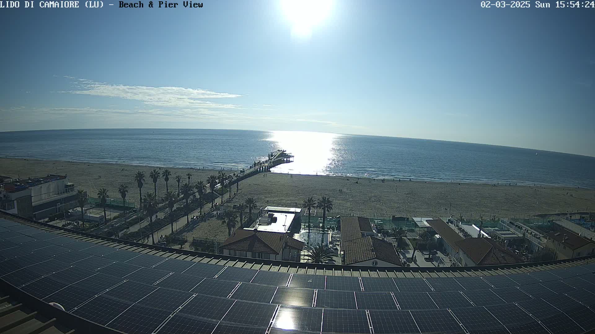 A sunny day overlooking a beach with a pier, palm trees, buildings, and a solar panel array on a rooftop.