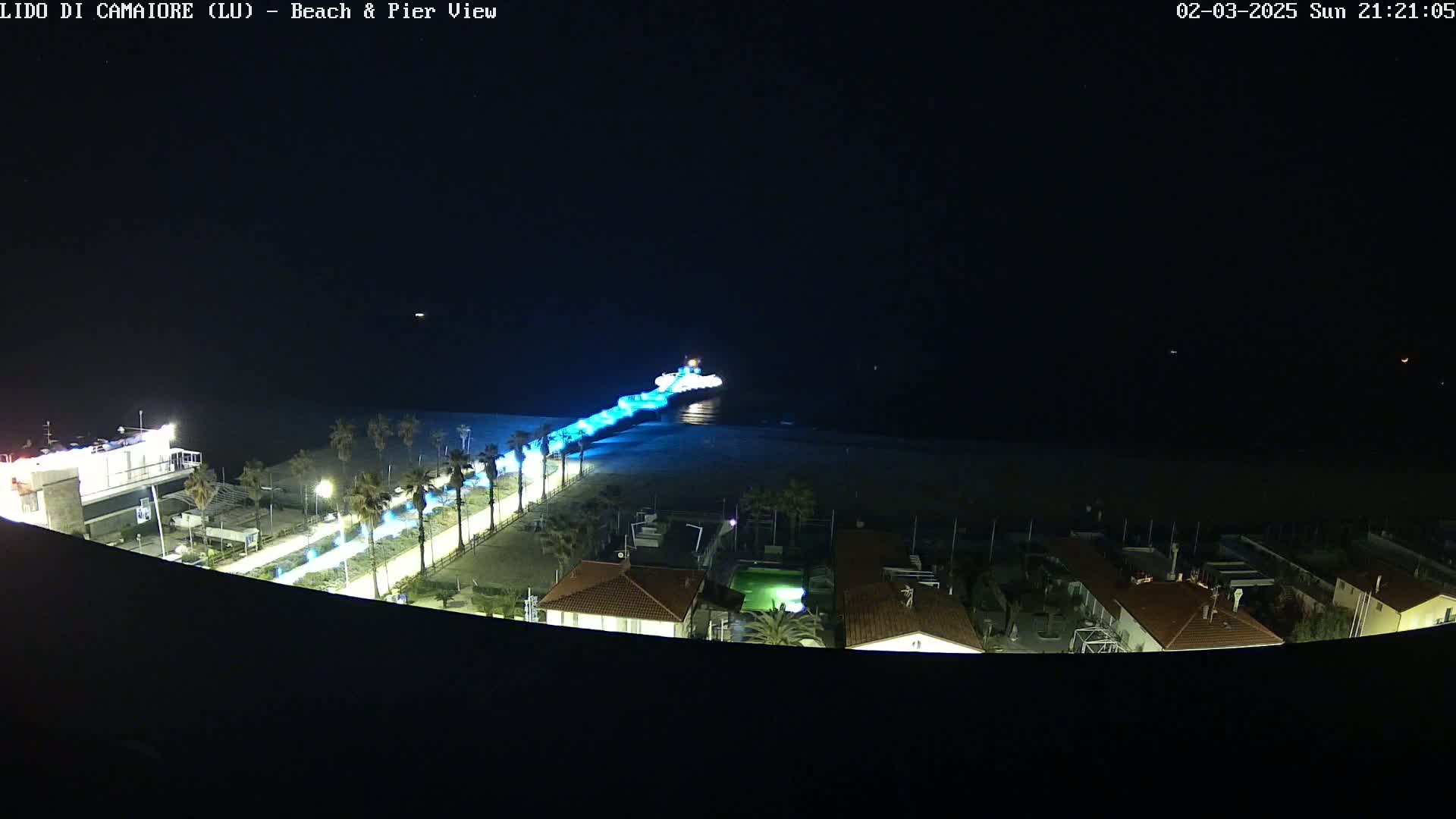 A nighttime aerial view shows a pier extending from a beach into the dark ocean, illuminated with blue lights, with palm trees and buildings visible along the shoreline.
