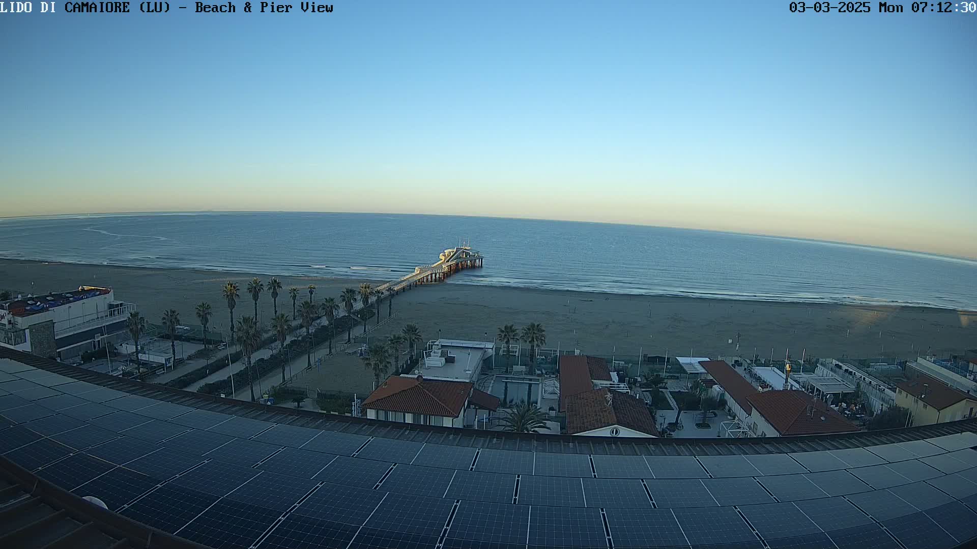 A pier extends into a calm sea under a clear, bright blue sky, overlooking a sandy beach lined with palm trees and buildings with solar panels on their roofs.