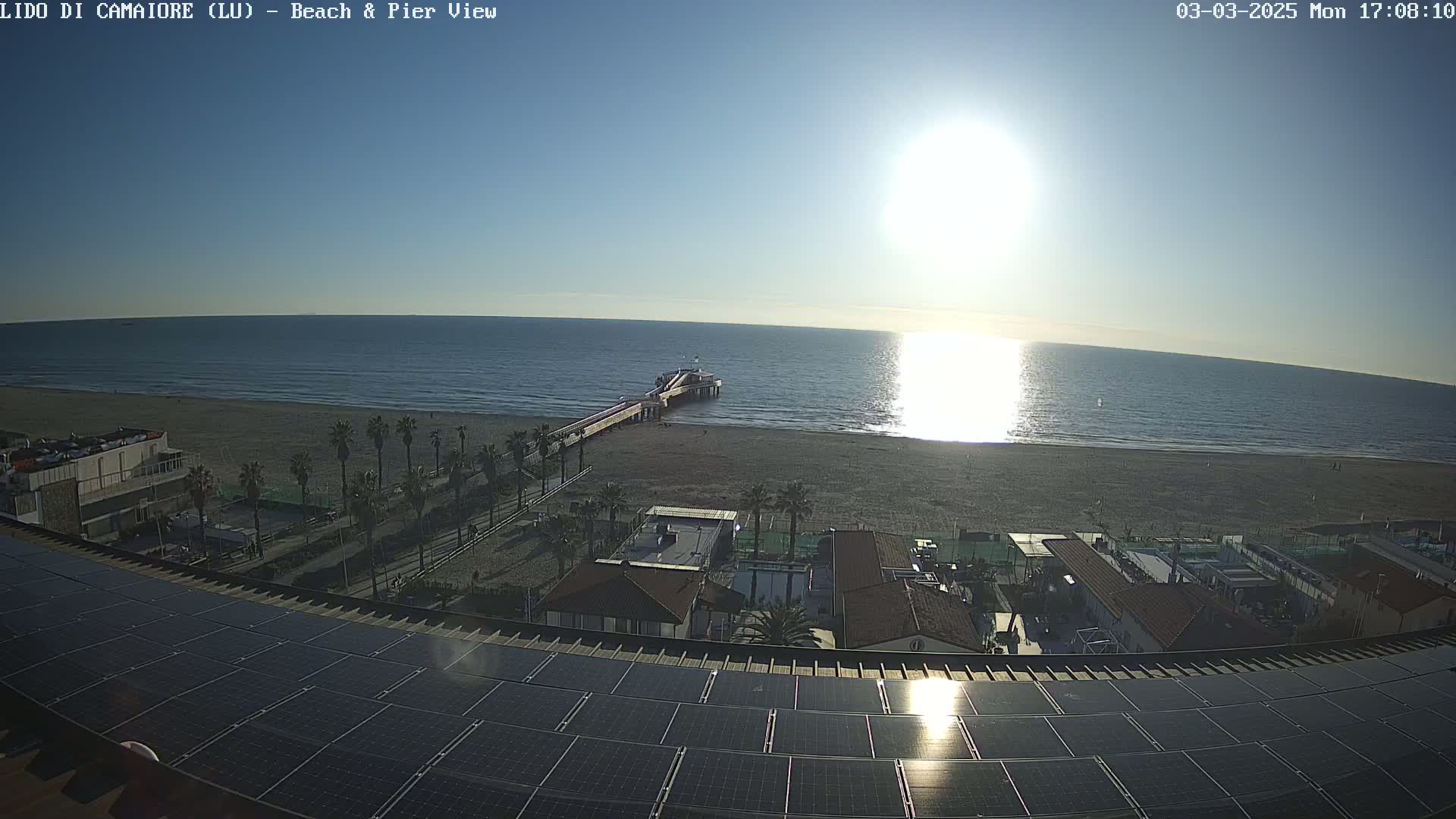 A sunny beach scene with a pier extending into a calm sea, viewed from a rooftop with solar panels.