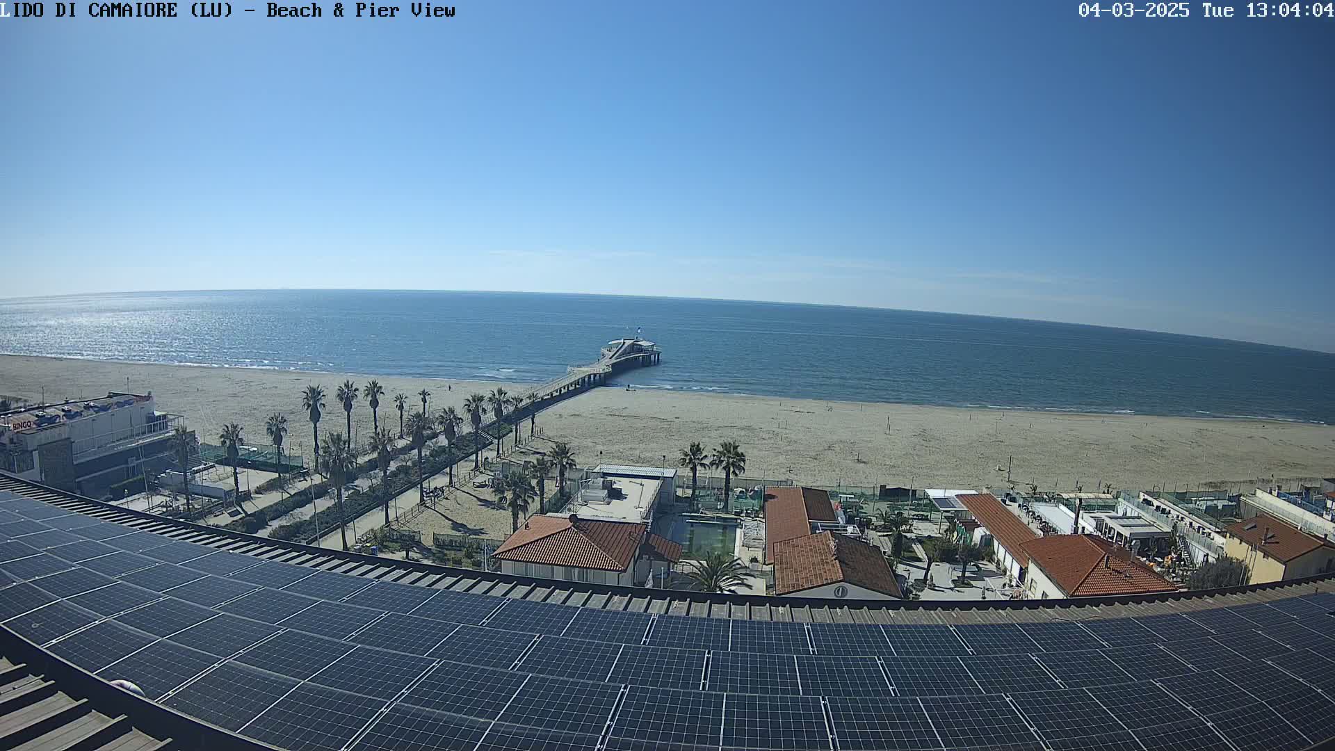 A sunny day shows a beach with a pier extending into a calm ocean, viewed from a rooftop covered in solar panels, with buildings and palm trees lining the beach.