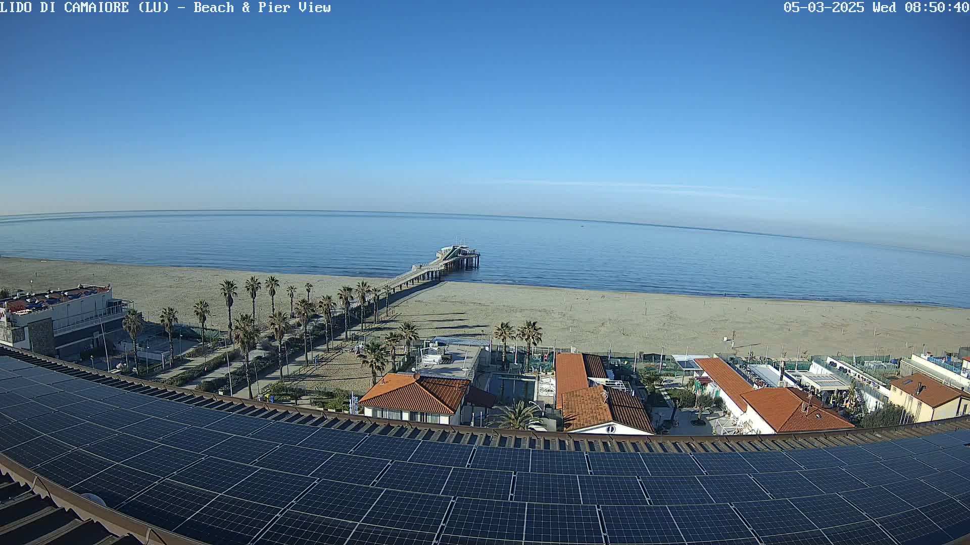 A beach scene on a sunny day shows a long pier extending into a calm ocean, with palm trees and buildings visible along the shore, all viewed from above a rooftop covered in solar panels.