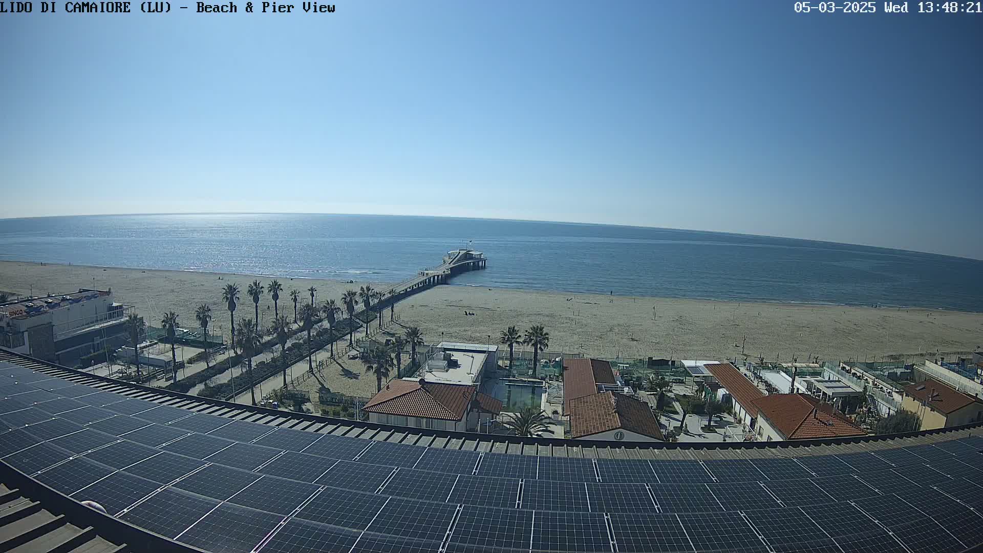 A sunny day at a beach shows a pier extending into a calm sea, with palm trees lining a sandy shore, all viewed from above a building equipped with solar panels.