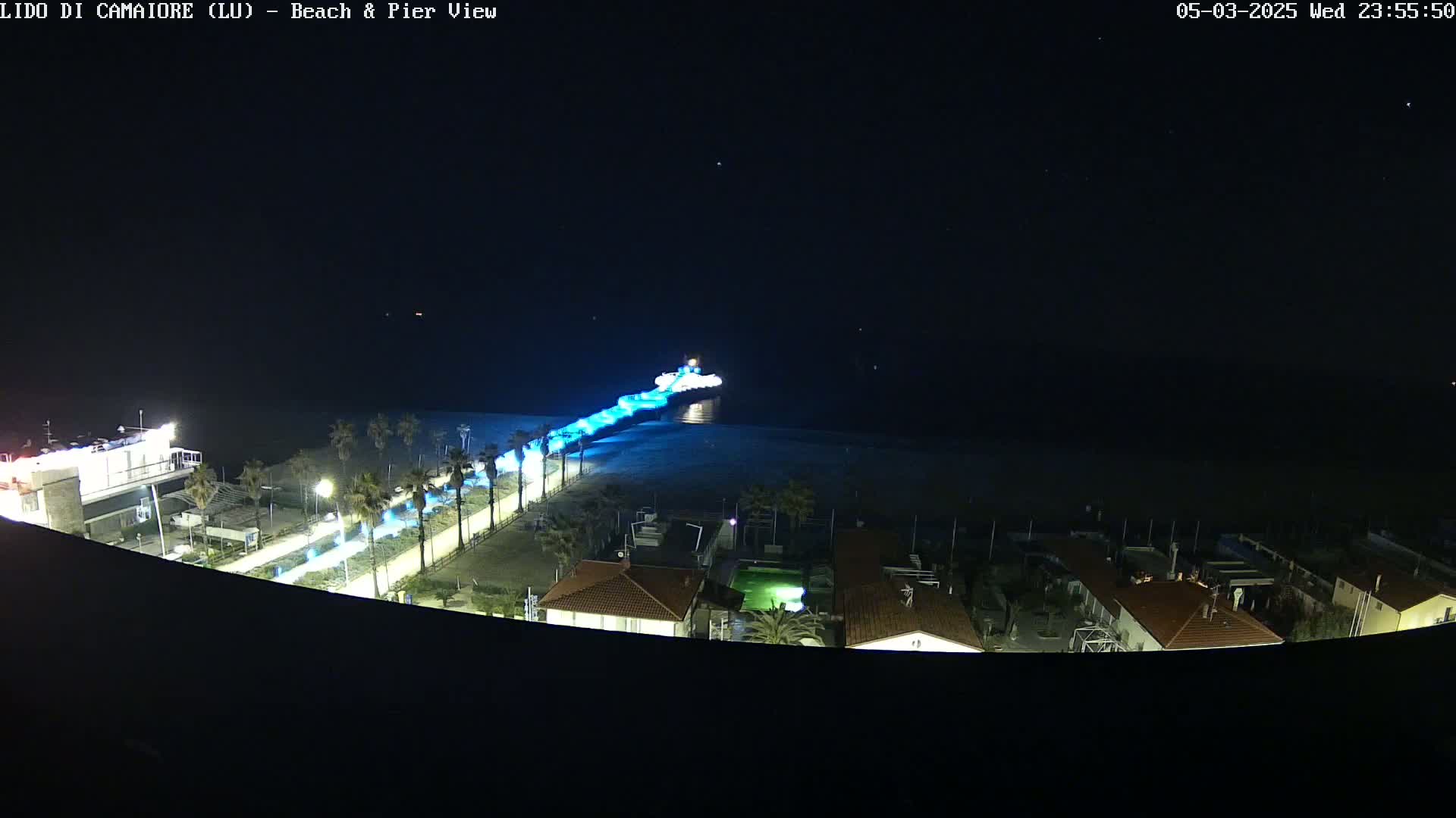 A nighttime, high-angle view shows a pier extending into the dark water, illuminated in blue, with a beach and buildings visible below.